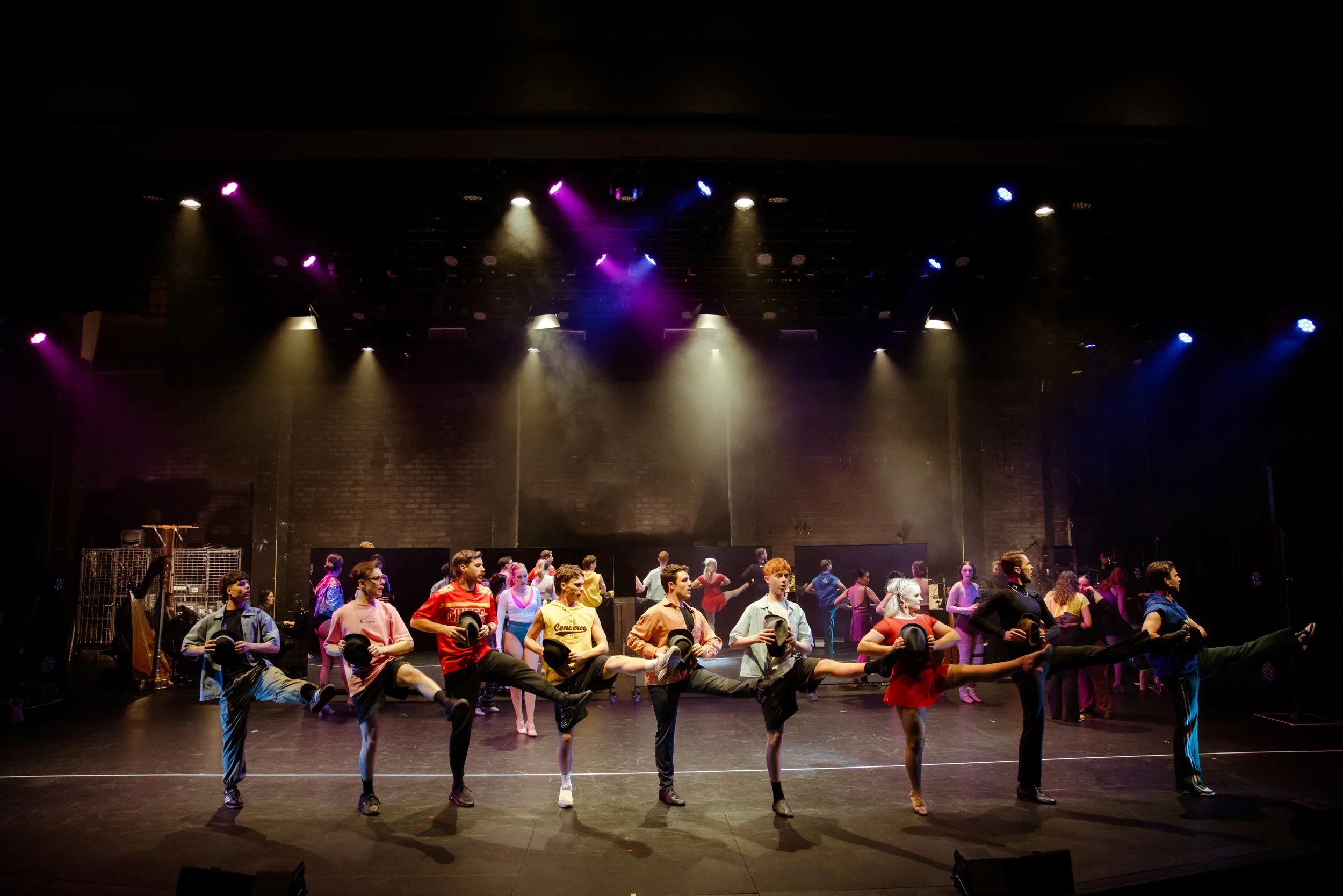 The full cast of A Chorus Line in a kick line under stage lights at Laycock Street Theatre