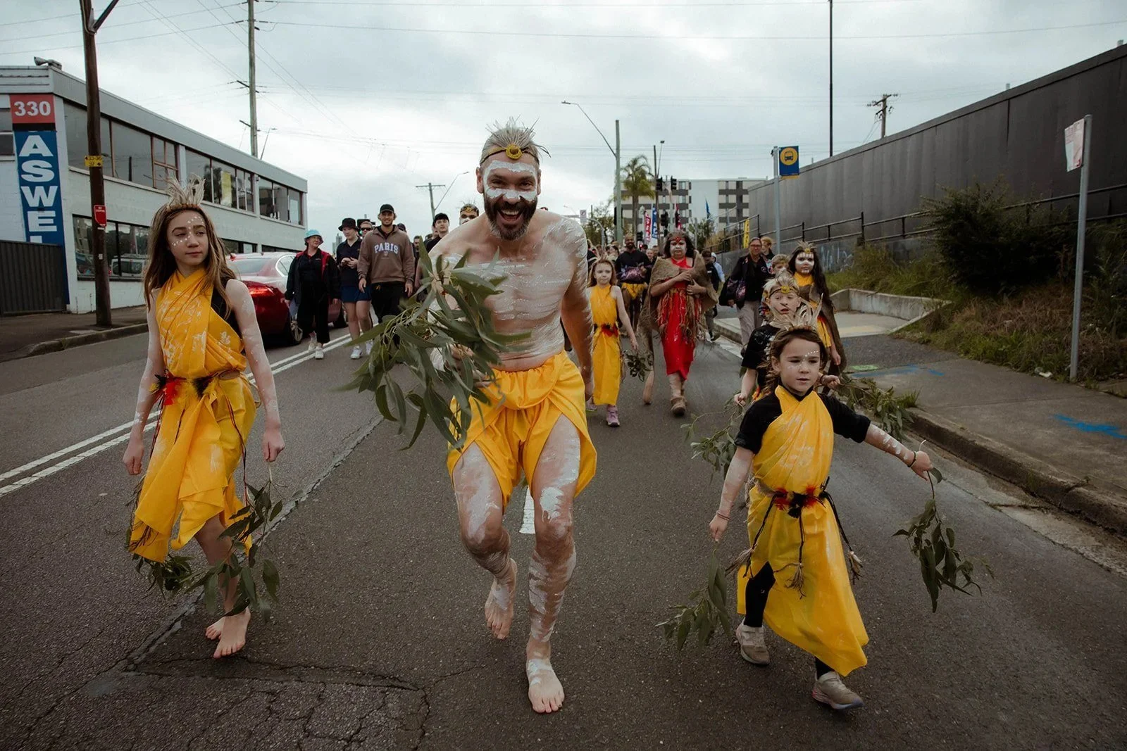 A painted dancer leads the Reconciliation Walk in Gosford, children in yellow following with gum leaves.