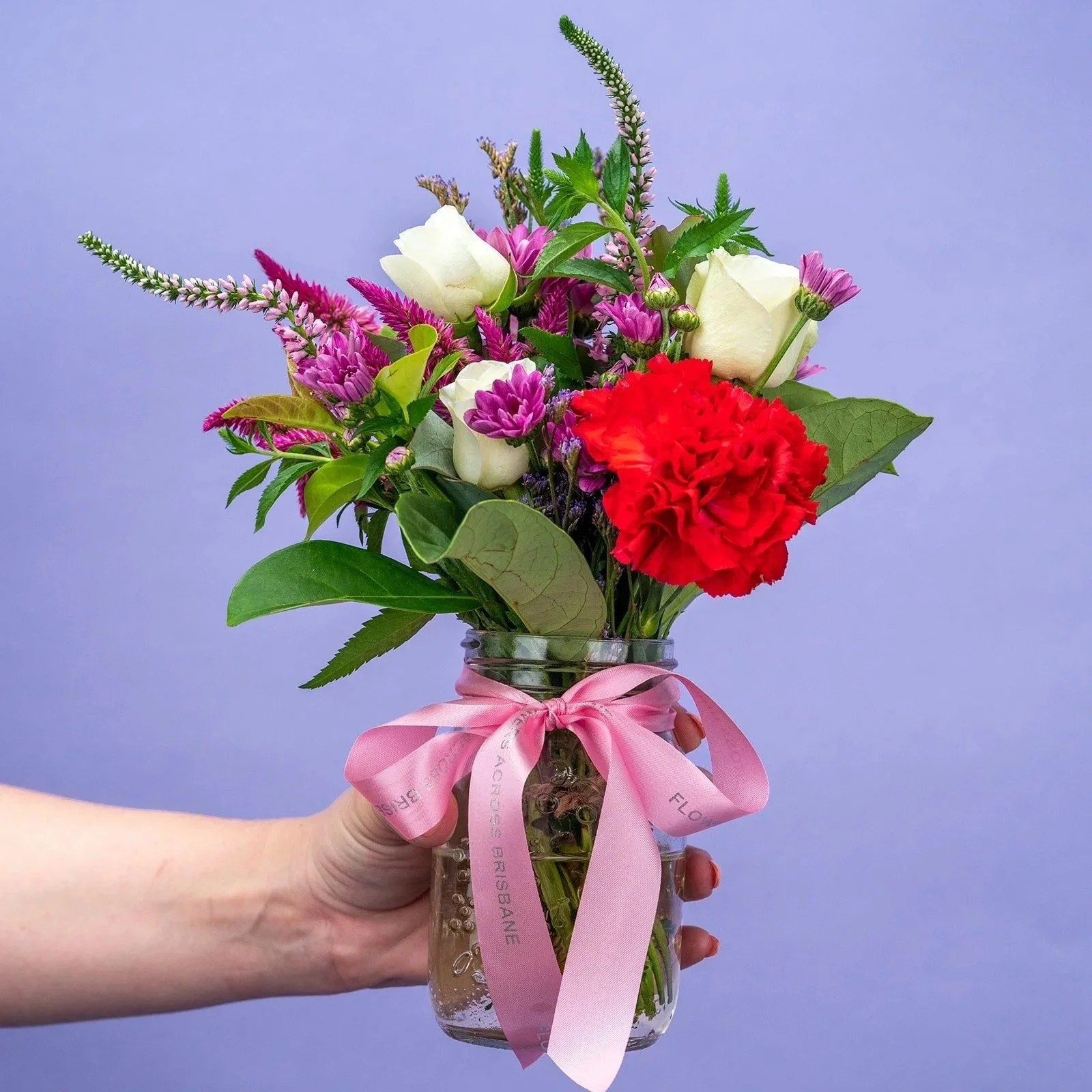 A hand holding a bouquet of red, pink and white flowers against a lilac backdrop