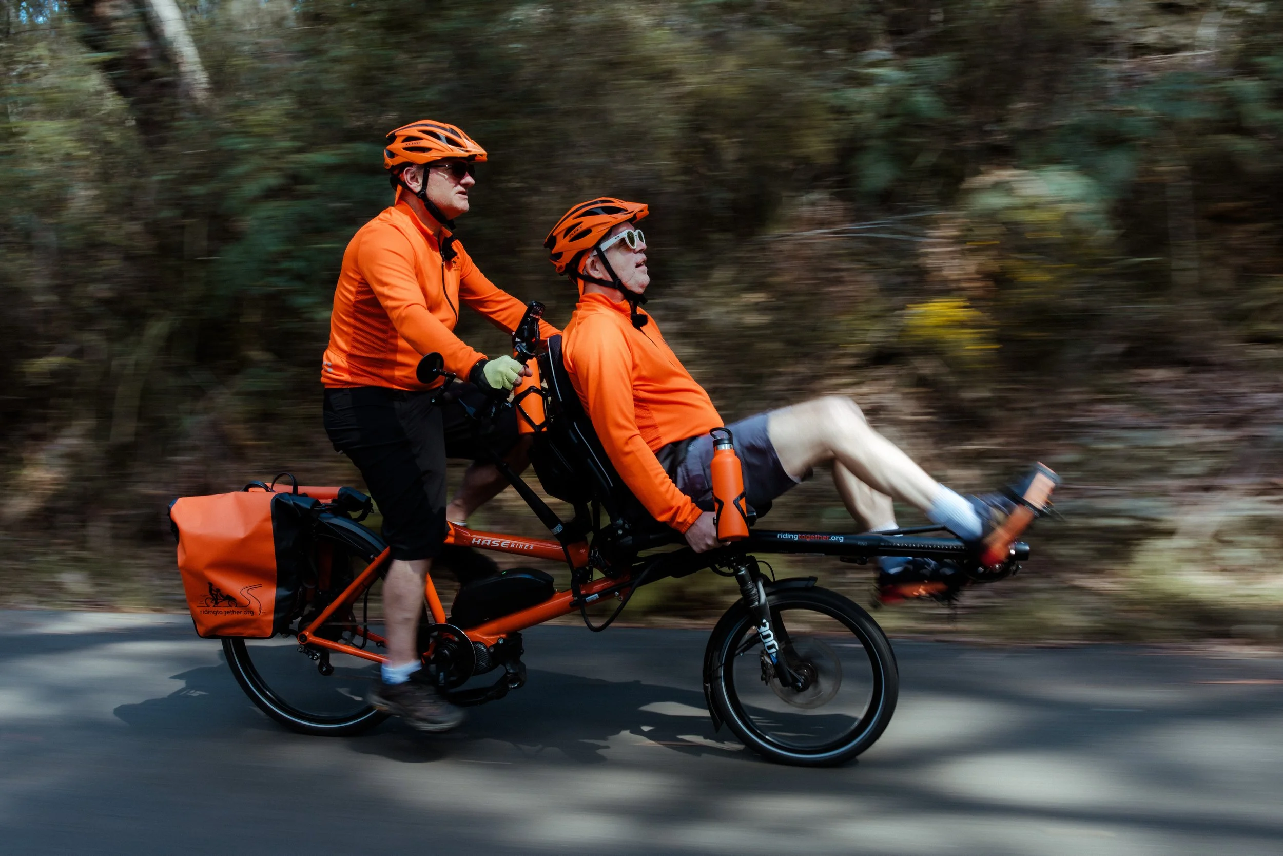 A Riding Together tandem e-bike at speed on a bush road, both riders in orange