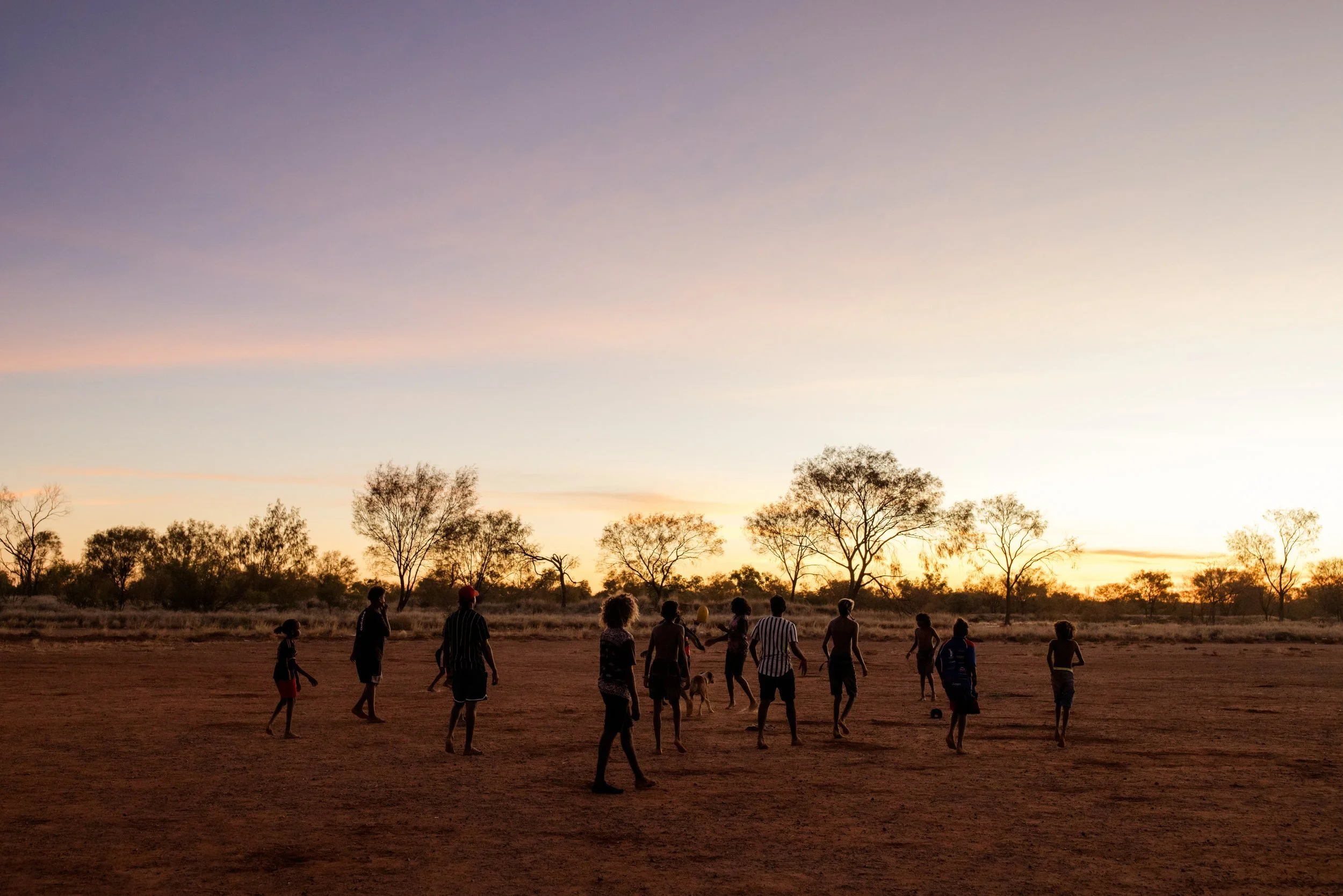 Silhouettes of players on a red dirt oval at dusk.