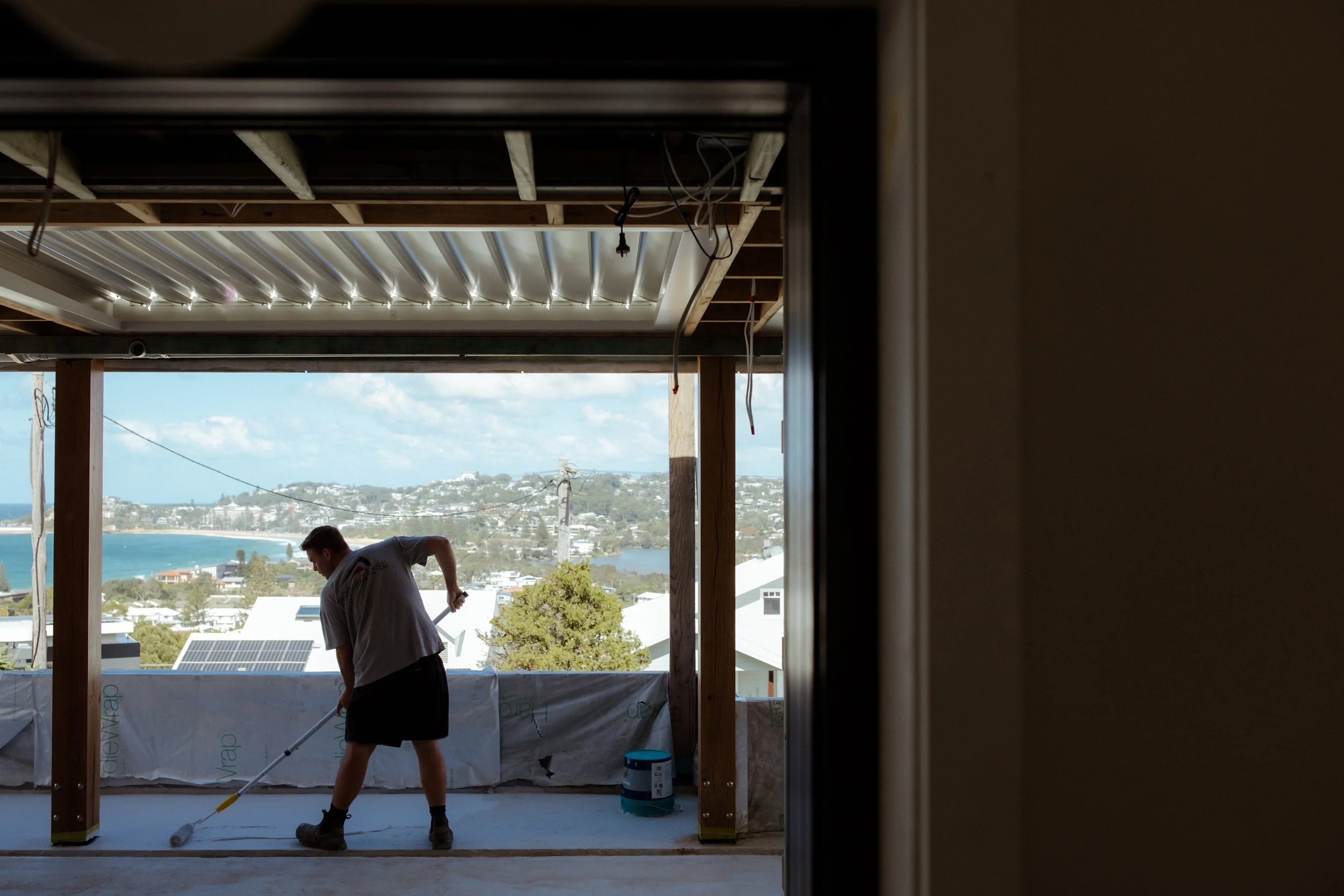 A worker rolls out deck membrane, framed by an open doorway with the Terrigal coast behind.