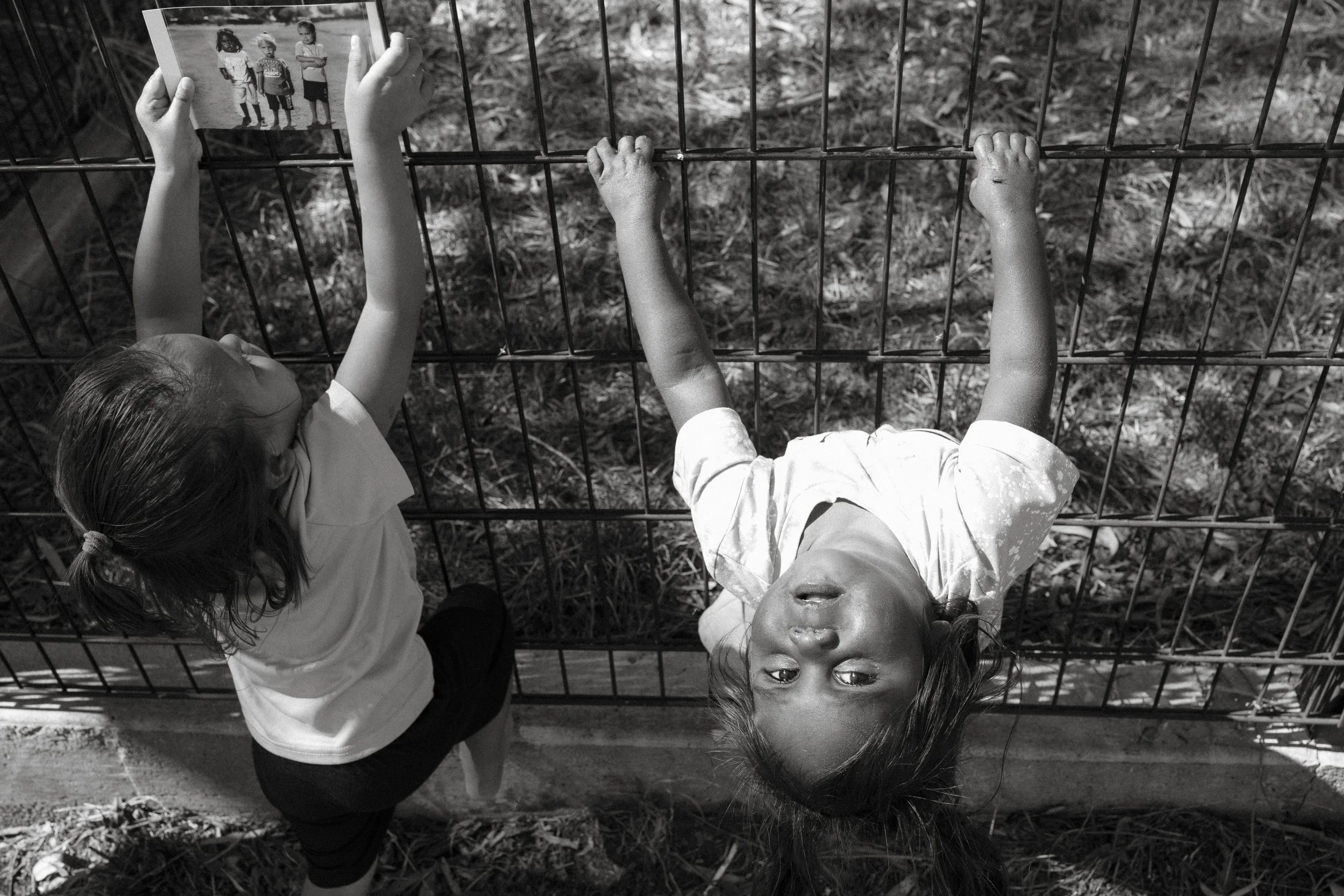 Two children hang upside down on a fence at Jigalong, one holding a print, in black and white.
