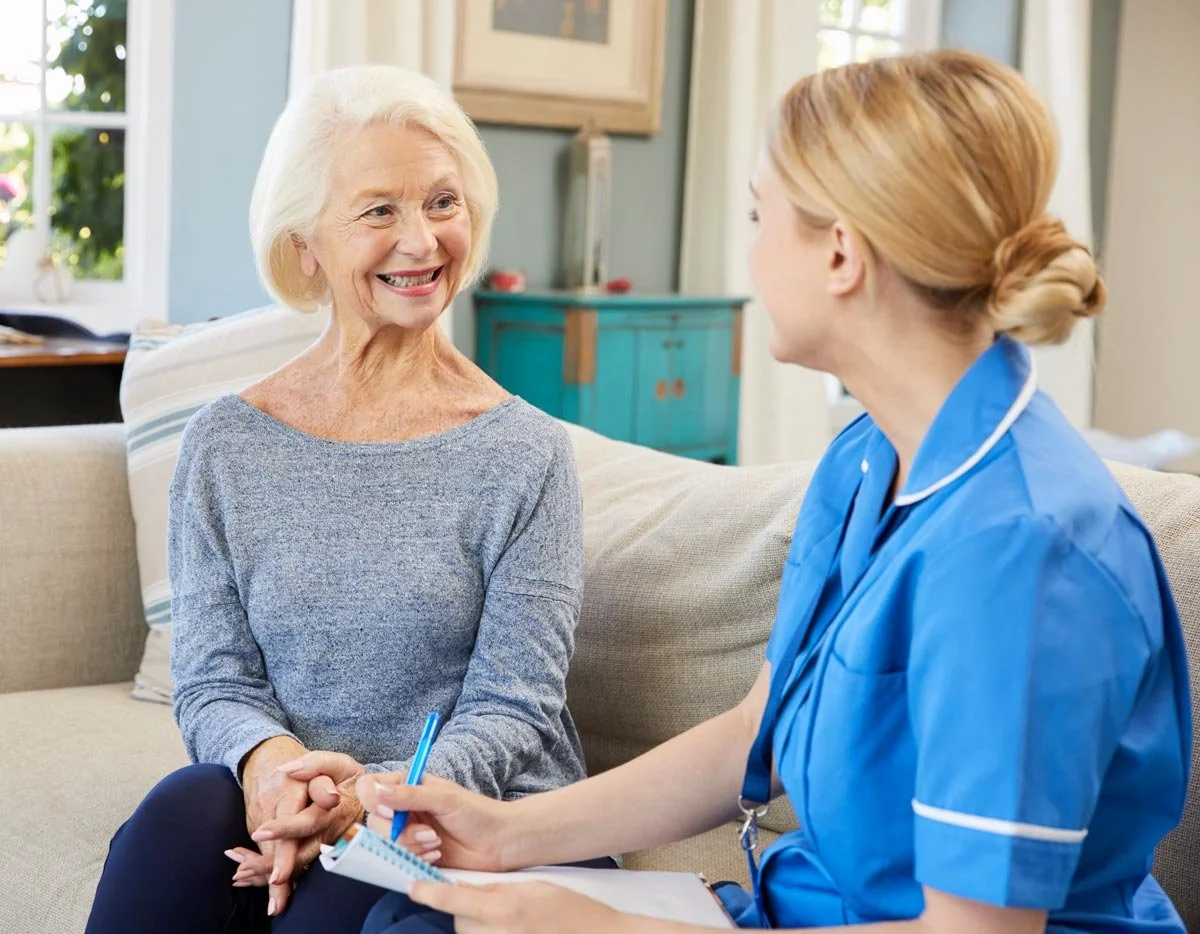 A woman sits on a couch, engaged in conversation with a nurse in a comfortable setting. | Feature image for the Fall Risk Assessments page