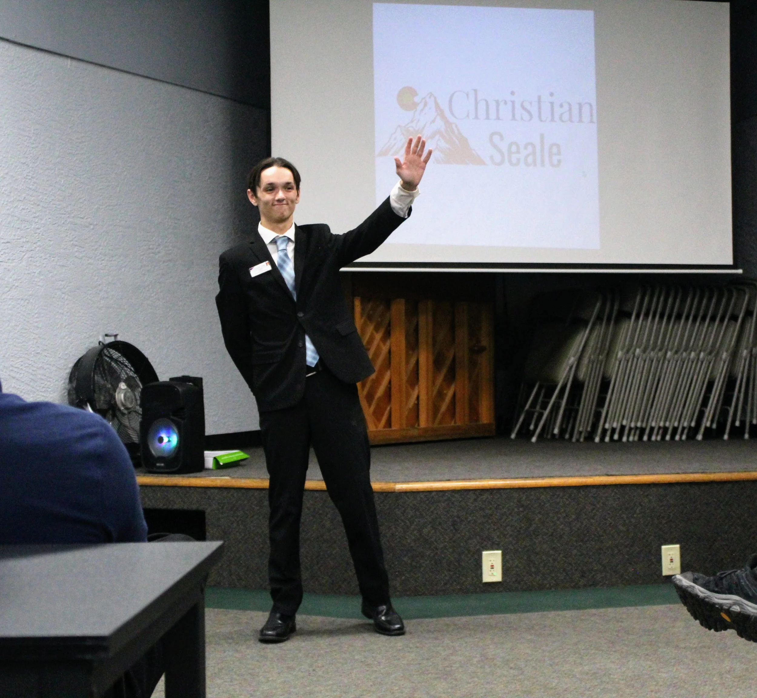 Christian Seale, candidate for El Paso County Commissioner District 5 standing on a small stage, smiling and waving at an audience. A large screen behind him displays a logo with a mountain, sun, and the text 'Christian Seale.' Several folded chairs 