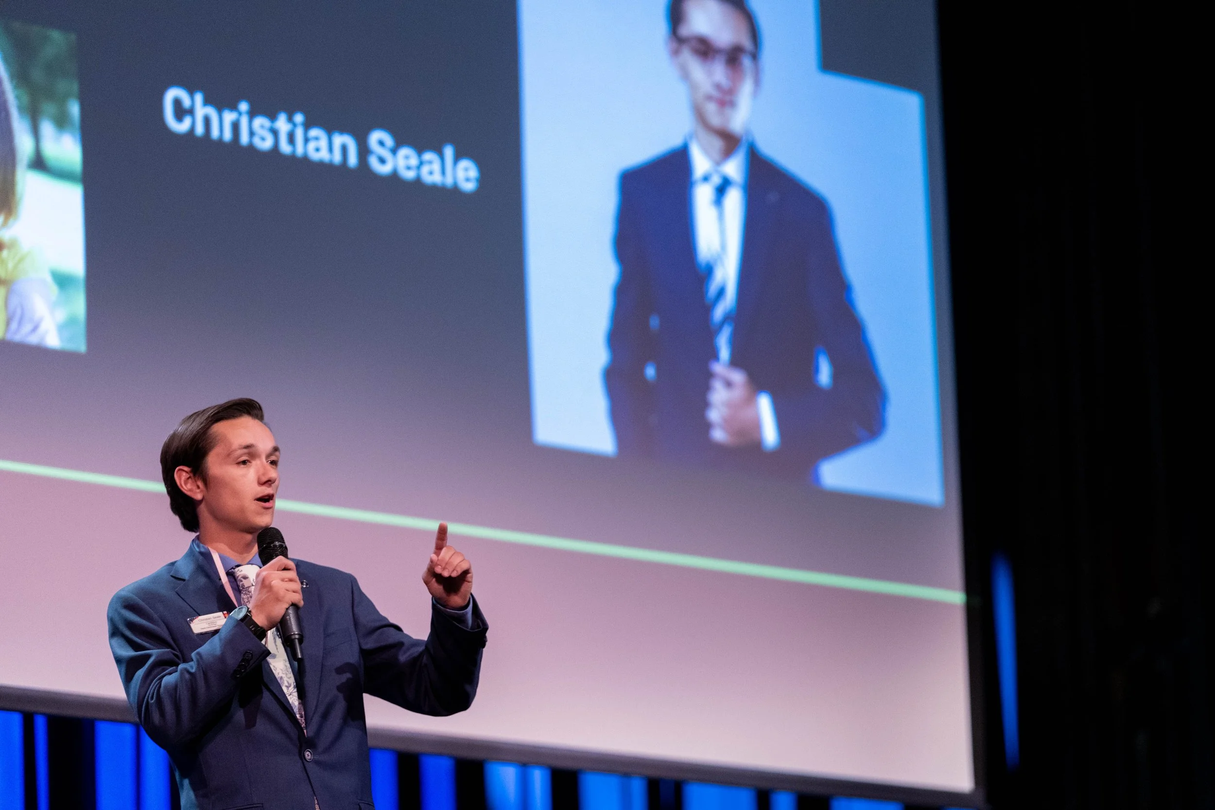 Christian Seale for County Commissioner District 5 in a suit is giving a presentation on stage, holding a microphone, with a large screen behind him displaying a slide that includes his photo and the name 'Christian Seale.'