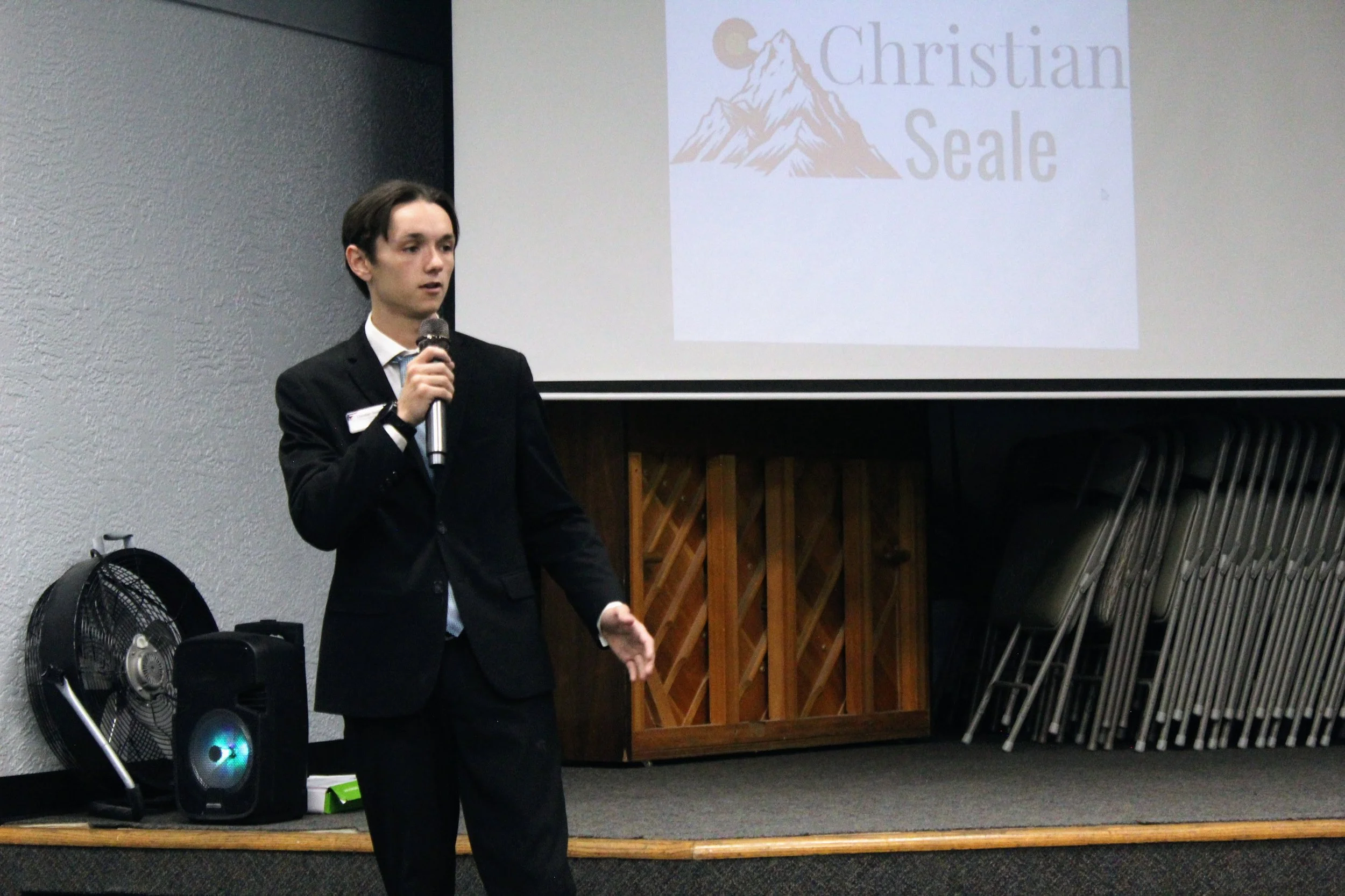 Christian Seale, candidate for El Paso County Commissioner District 5 giving a presentation with a microphone in front of a slide that reads 'Christian Seale' and features a mountain and moon logo.