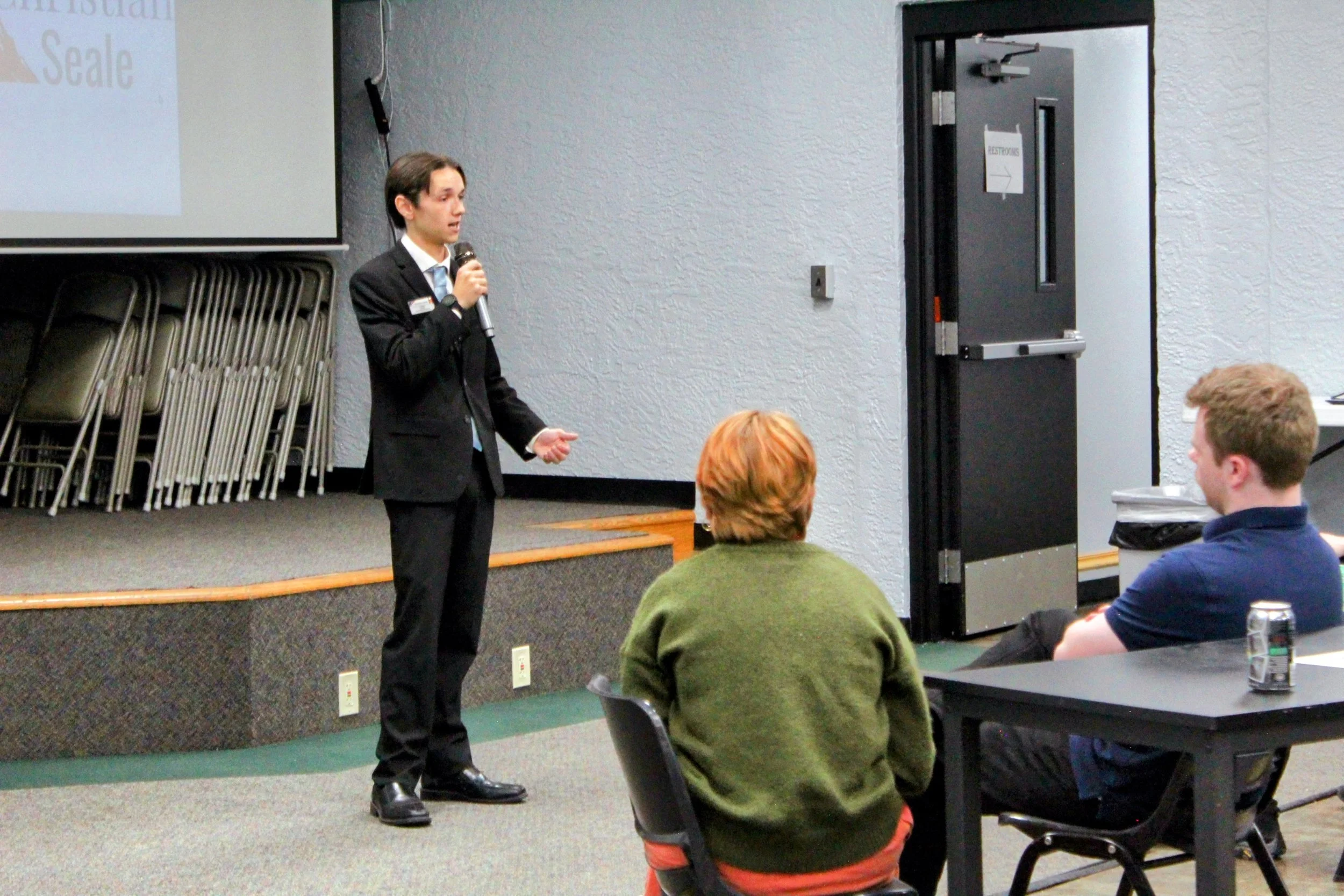 Christian Seale, candidate for El Paso County Commissioner District 5 presenting with a microphone in a conference room, facing an audience.