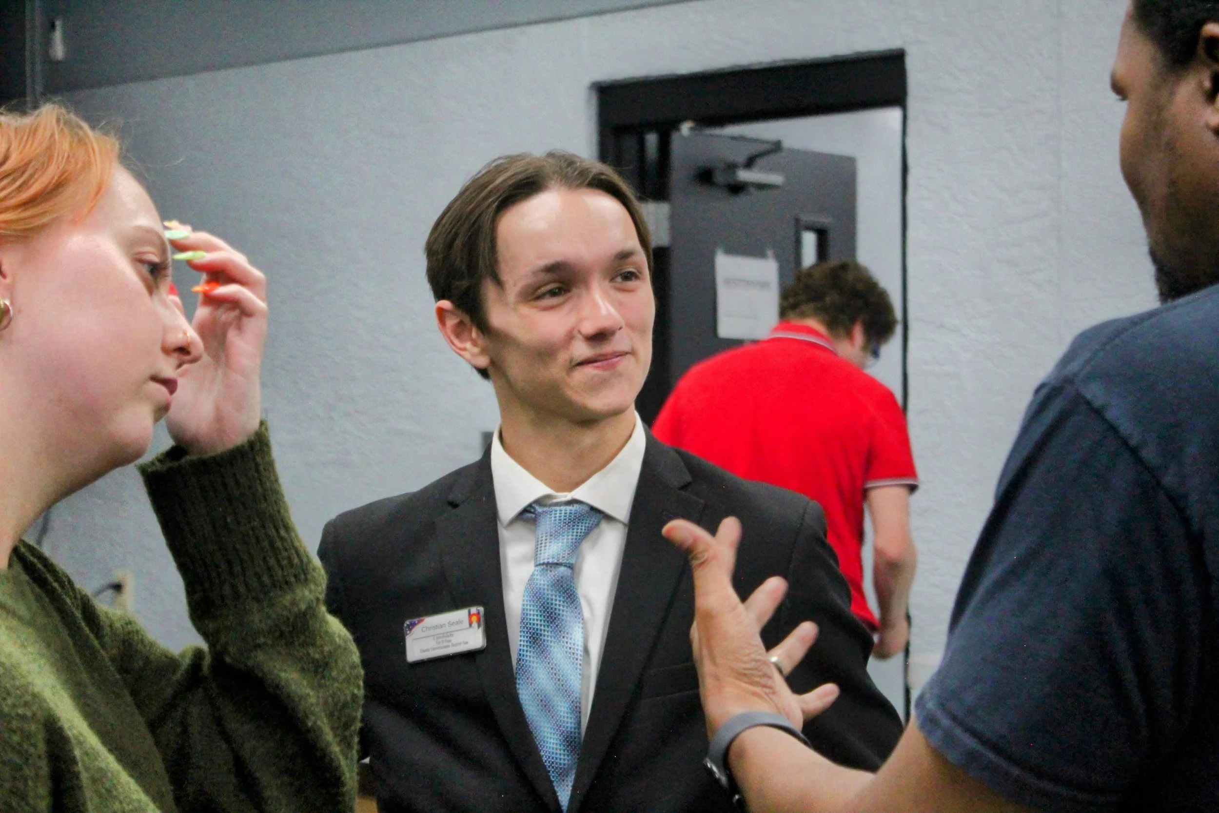 Three people engaged in a conversation indoors, with a woman on the left adjusting her hair, Christian Seale, candidate for El Paso County Commissioner District 5 in the center smiling and wearing a suit with a name badge, and a man on the right gest