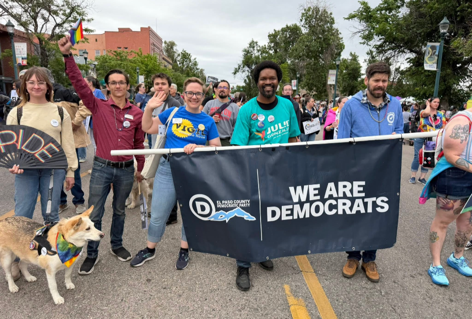 Christian Seale candidate for el paso with a group of diverse people participating in a pride parade, holding a banner that reads "WE ARE DEMOCRATS" and displaying Democratic Party symbols, with some waving flags and others smiling at the camera.