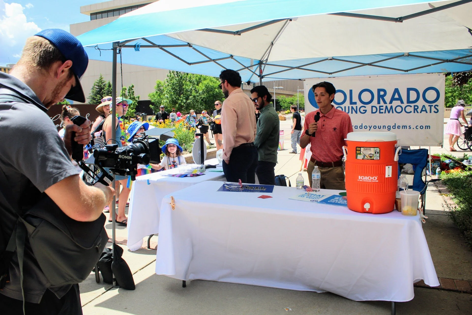 Christian Seale, candidate for El Paso County Commissioner District 5, promoting the Colorado Young Democrats. There is a person speaking into a microphone, and a camera operator recording the event. Children and other attendees are visible in the ba