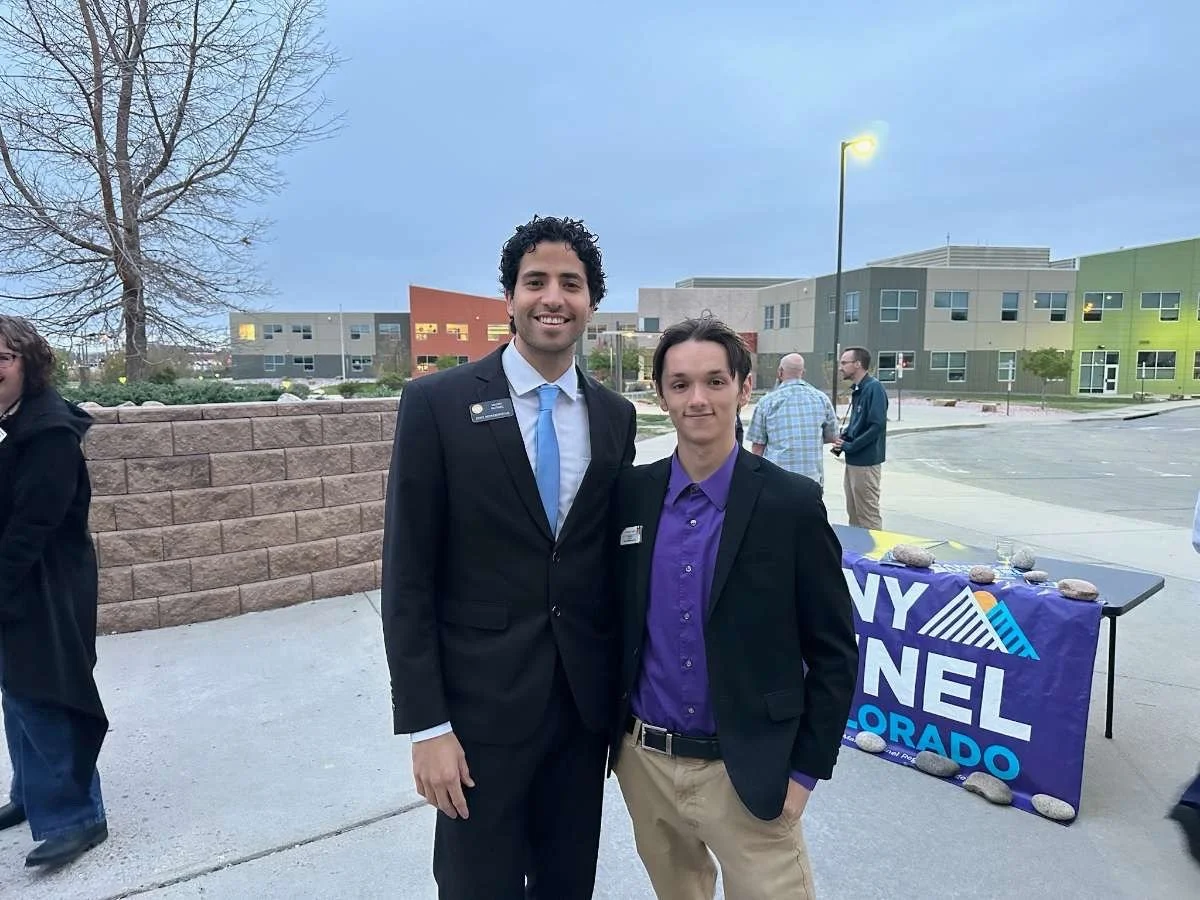 Christian Seale, candidate for El Paso County Commissioner District 5 and manny rutinel smiling at an outdoor event, with a political campaign table in the background and a colorful modern building behind them.