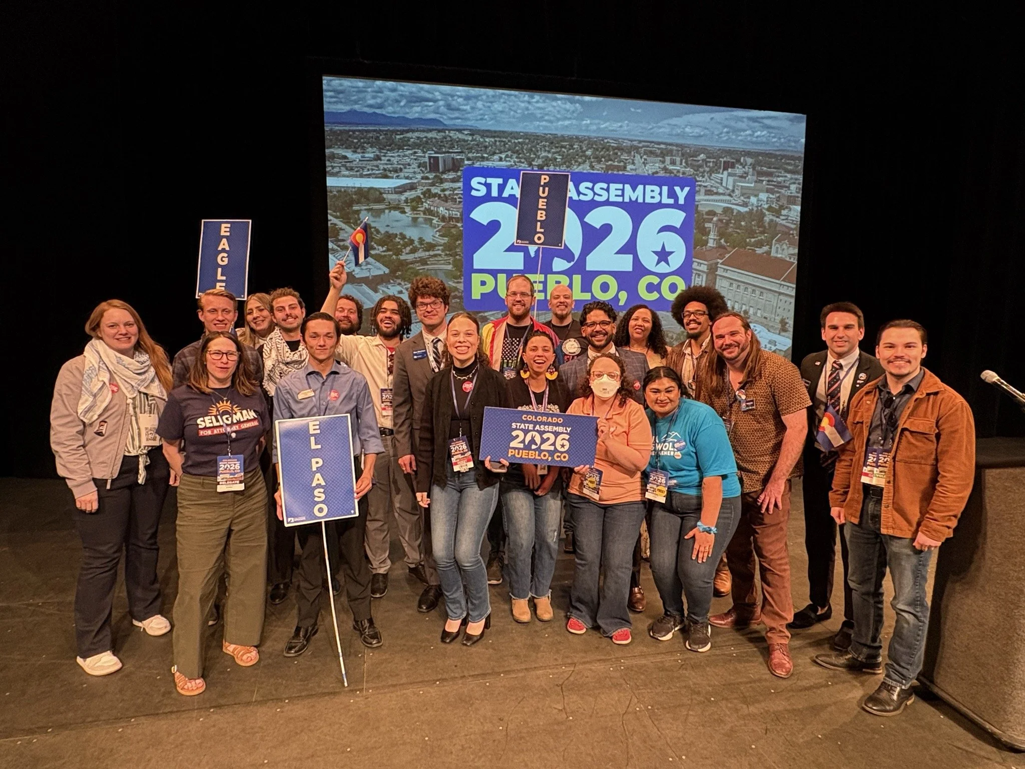 Group of diverse people gathered on stage, holding signs and flags, celebrating at the Colorado State Assembly 2026 in Pueblo, Colorado.