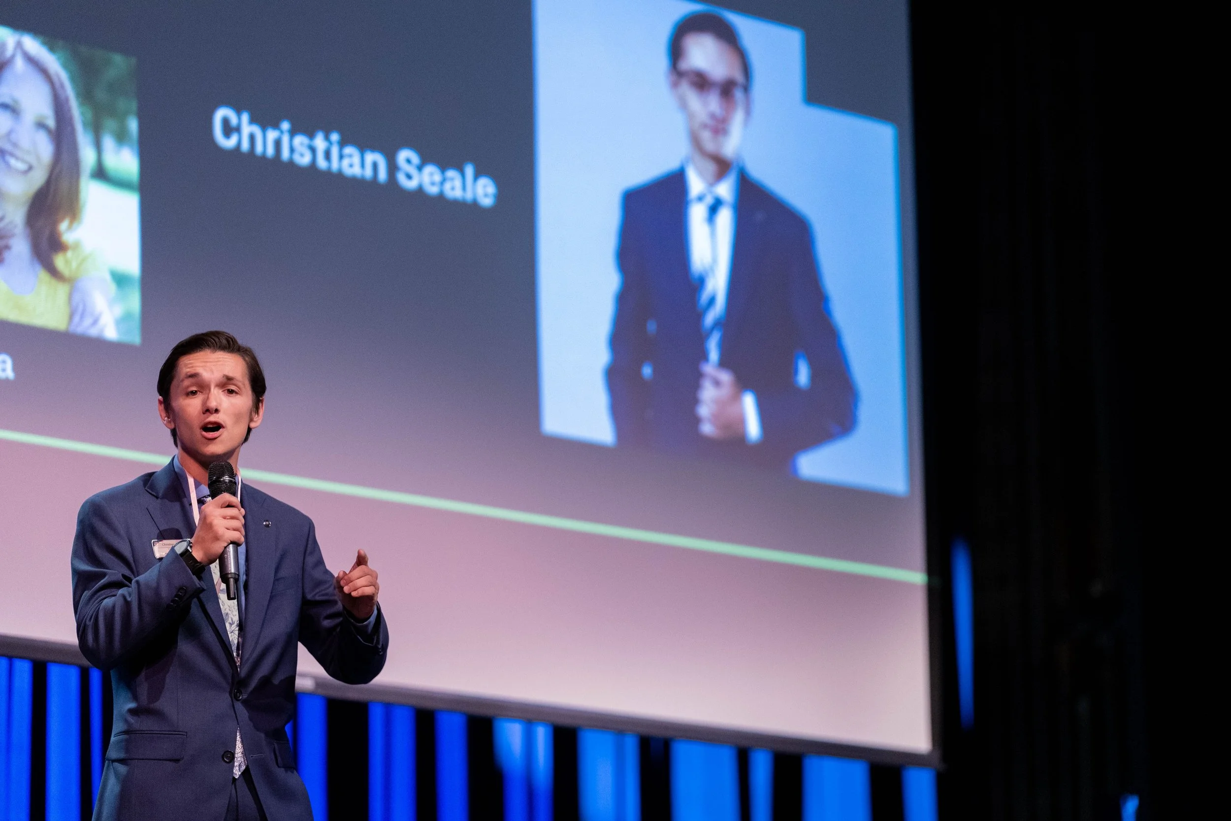 Young man in a blue suit speaking into a microphone on stage, with a large screen behind him displaying his photo and the name 'Christian Seale'.