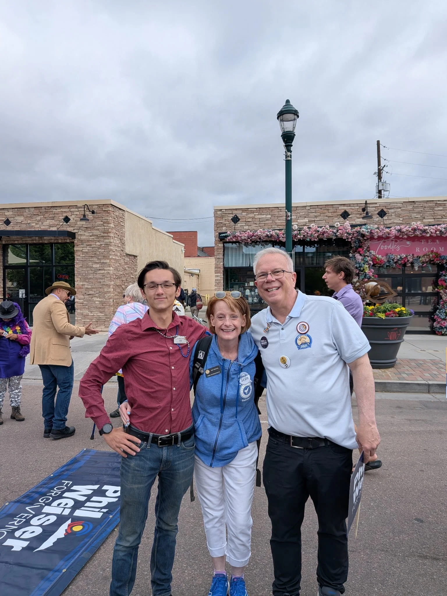 Christian Seale candidate for el paso county commissioner district 5 with 2 others in front of a brick storefront decorated with pink flowers, with a lamp post and other pedestrians in the background.