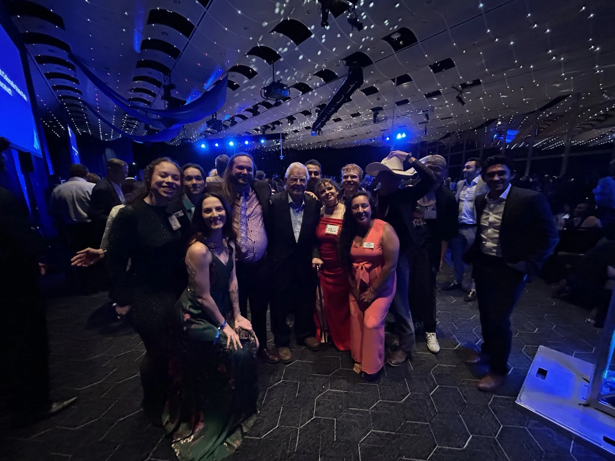 Group of people with Christian Seale, candidate for El Paso County Commissioner District 5 dressed in formal attire, posing together in a dark, illuminated event hall with blue lighting, ceiling decorations, and some seated audience members in the ba
