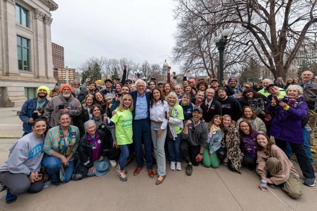 Group of diverse people with bernie sanders and AOC and Christian Seale, candidate for El Paso County Commissioner District 5 gathered outdoors on a city street, smiling at the camera, with the Capitol dome visible in the background.