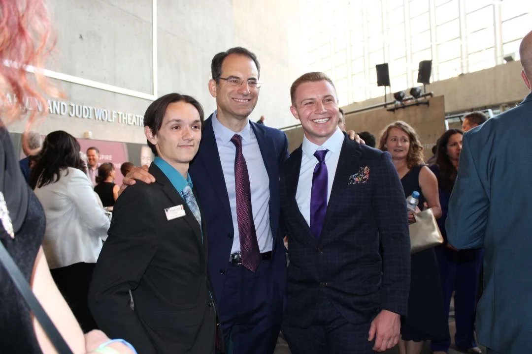 Christian Seale, candidate for El Paso County Commissioner District 5 and Phil Wisner, smiling and posing together at an indoor event with several other people in the background.