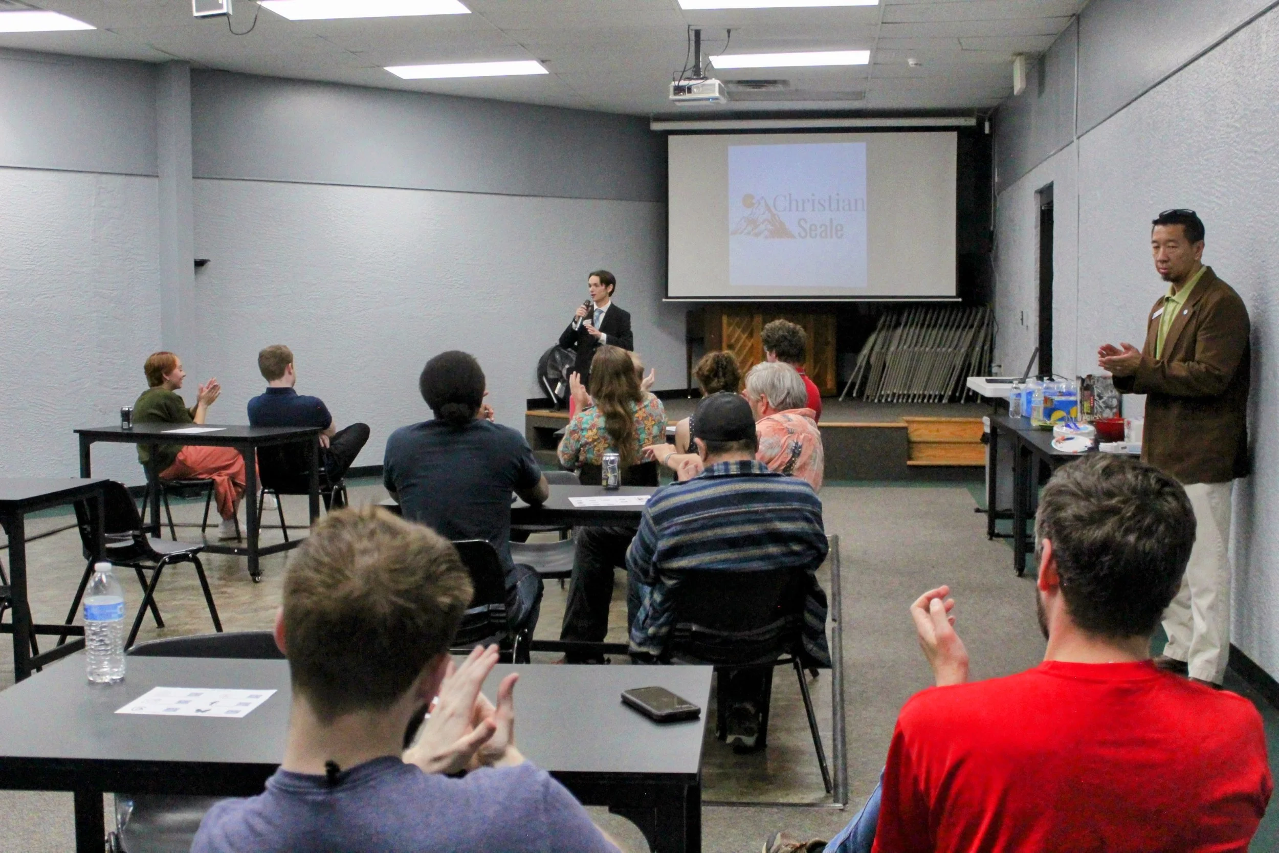 Christian Seale, candidate for El Paso County Commissioner District 5 speaking at a presentation in a room with an audience. There's a large screen displaying the name Christian Seale, with an image of mountains and a sunset. Others are sitting and l