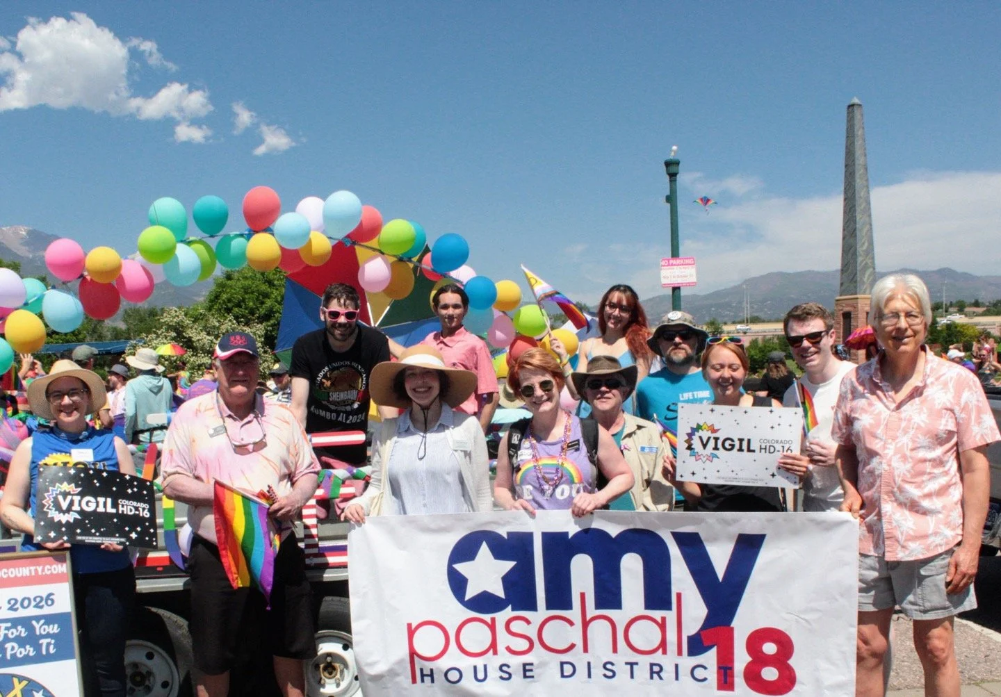 Group of people with Christian Seale, candidate for El Paso County Commissioner District 5 holding signs and rainbow flags at a Pride parade, with balloons, colorful decorations, and mountains in the background.
