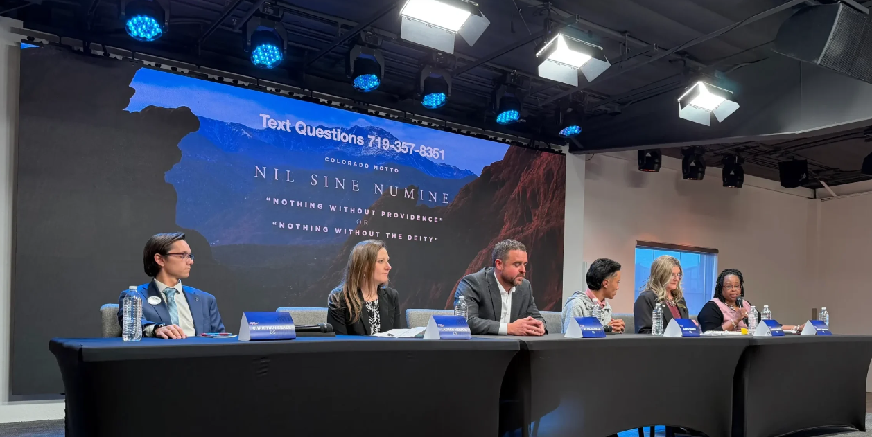 Christian Seale and 5 other people sitting at a long table on a stage, at Chruch of all Nation County Commissioner Froum. A large screen behind them displays a mountain landscape, a Colorado motto, and a phone number for text questions.