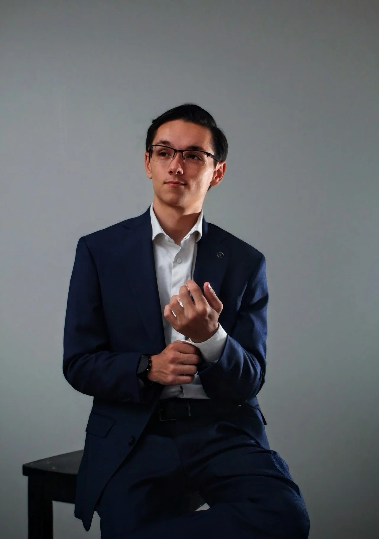 A young man in a navy suit and white shirt, wearing glasses, sitting on a black table against a plain gray wall, looking thoughtfully to the side.
