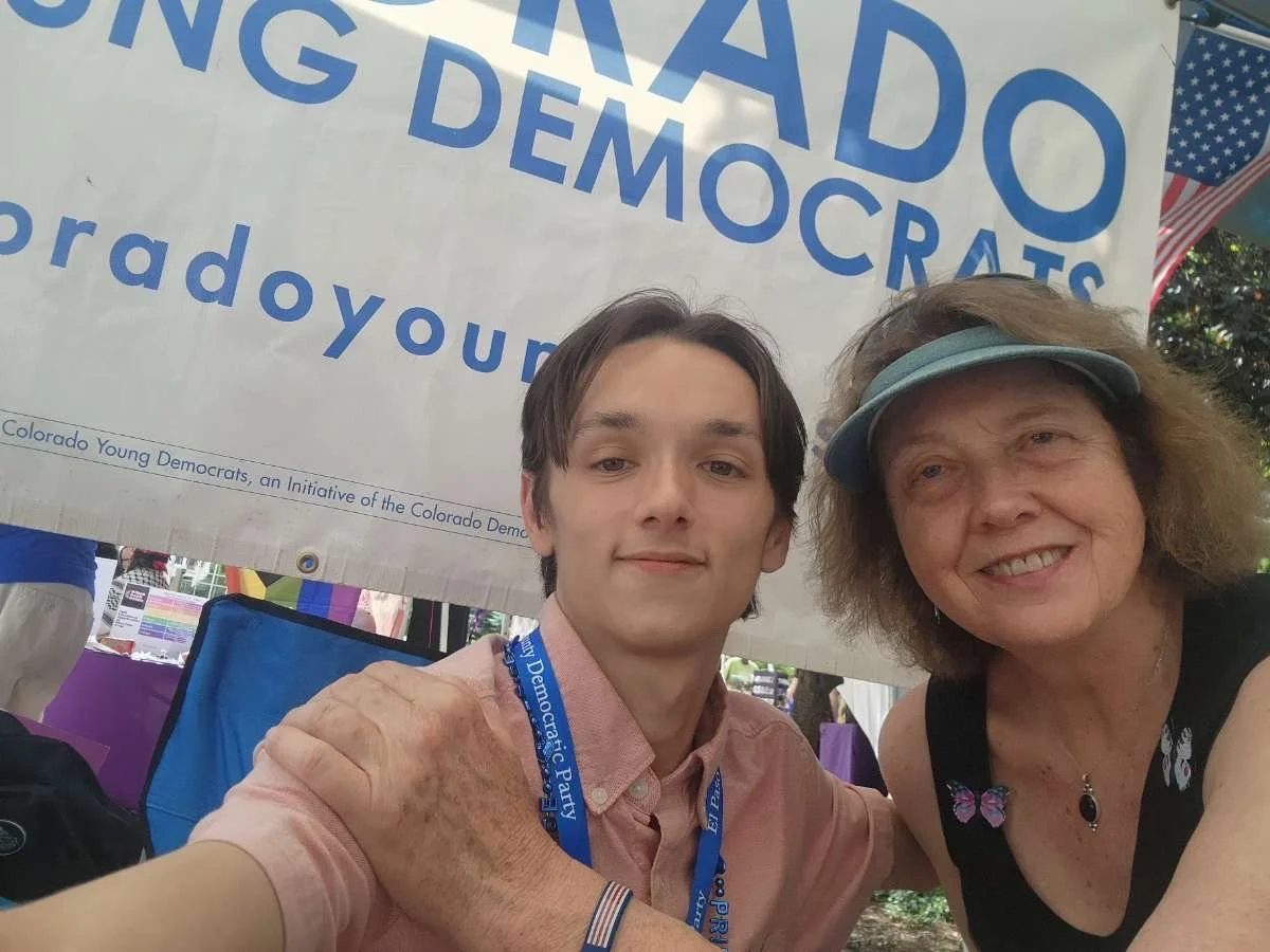 Christian Seale, candidate for El Paso County Commissioner District 5 taking a selfie in front of a United States flag and a banner that reads 'Colorado Young Democrats.' The person on the left is a young man with brown hair, wearing a pink shirt and
