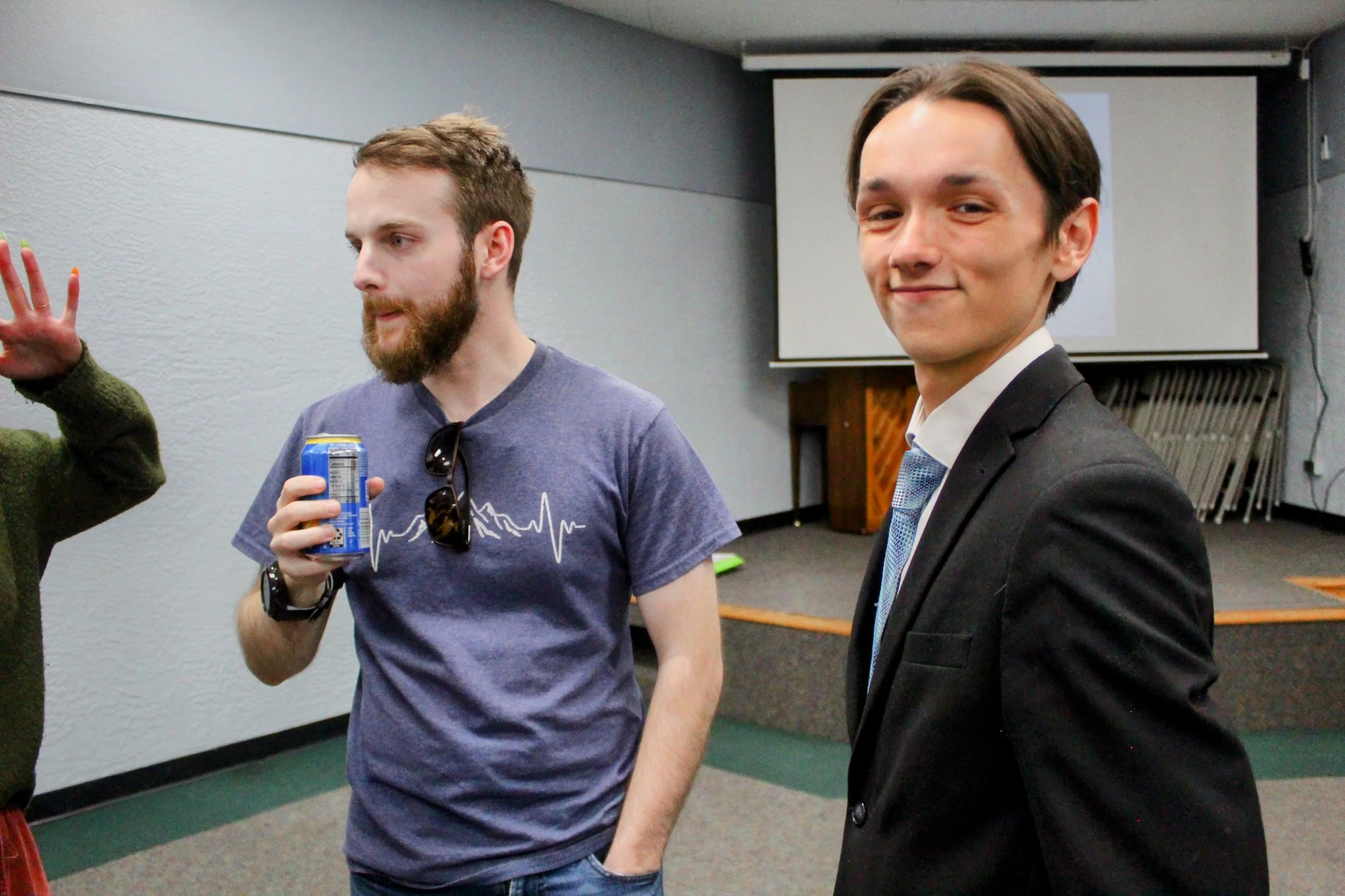 Christian Seale, candidate for El Paso County Commissioner District 5 standing in a room, one holding a soda can and wearing sunglasses on his shirt, the other in a suit and tie, smiling at the camera, with a projection screen in the background.