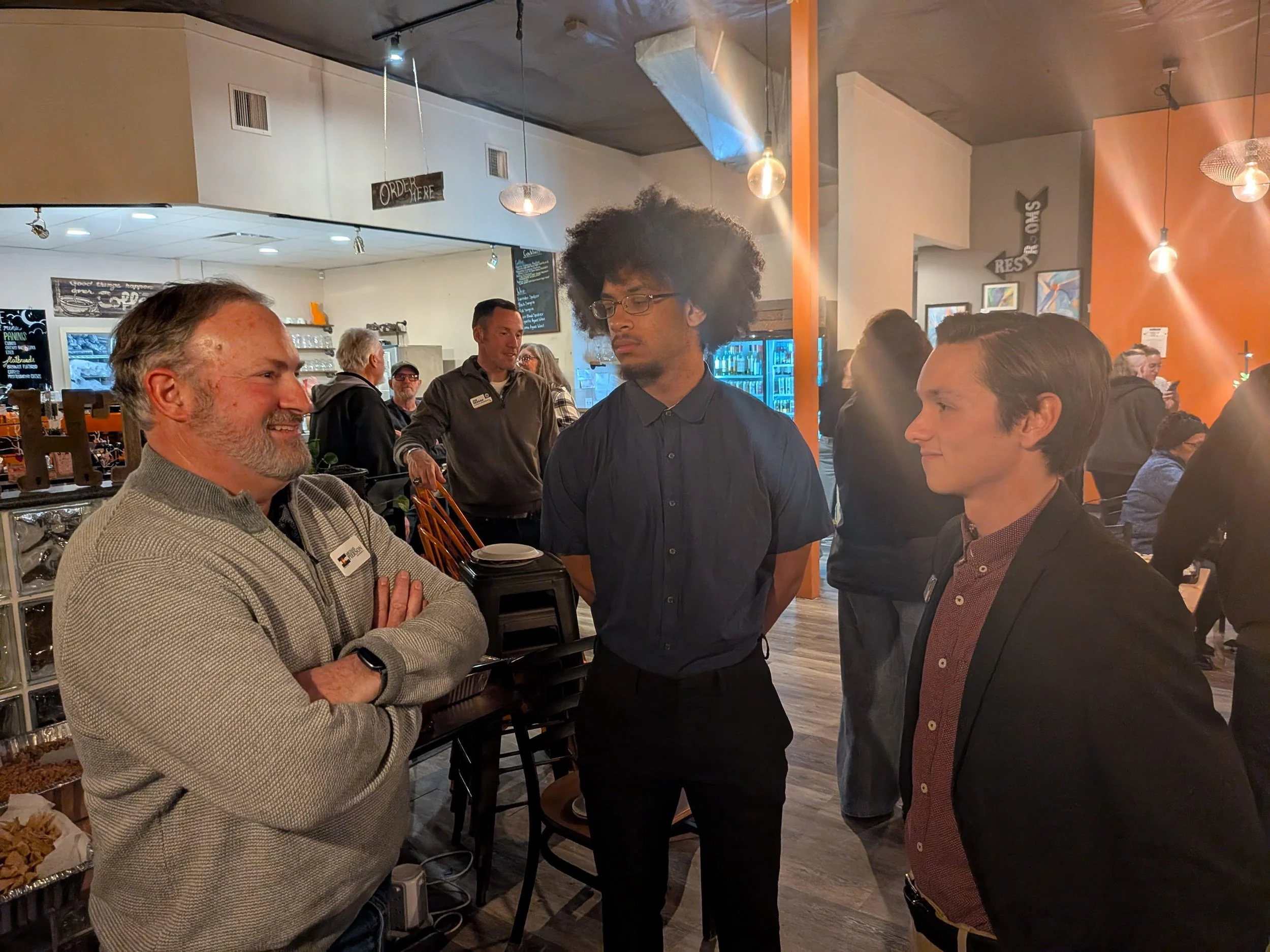 Christian Seale for County Commissioner are engaged in conversation inside a busy restaurant or café. Two of them are smiling, while the third appears more serious. The background shows other patrons and staff, with warm lighting and wooden accents.