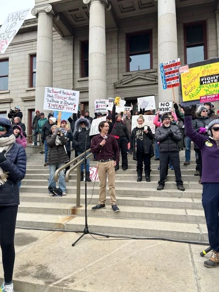 Crowd of protesters holding signs, some with messages like 'Make America Think Again' and 'Hands Off Our Democracy,' gathered on the steps of a government building with columns. Christian Seale, candidate for El Paso County Commissioner District 5 sp