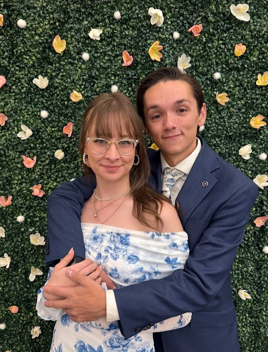 Talena Gipson and Christian Seale, candidate for El Paso County Commissioner District 5 standing close together, smiling, in front of a green leafy background decorated with flowers, wearing formal attire.