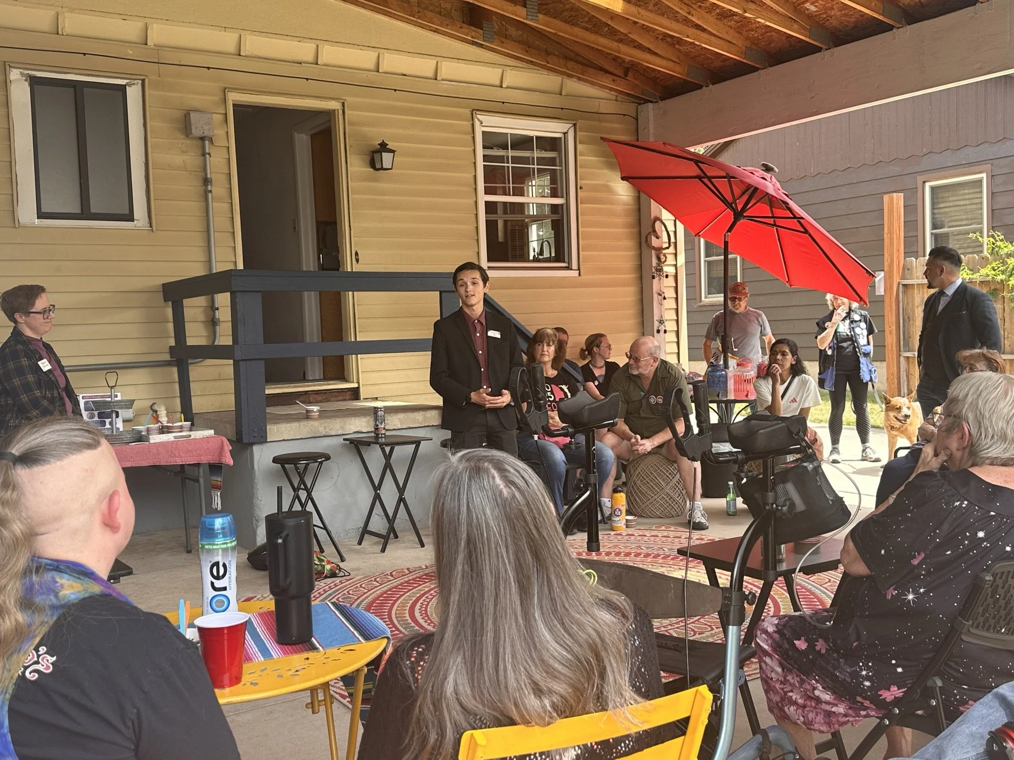 People outdoors on a porch with Christian Seale candidate for el paso county commissioner district 5 during a house party. There is a red umbrella, various chairs, and a table with drinks, with a yellow house and fences in the background.
