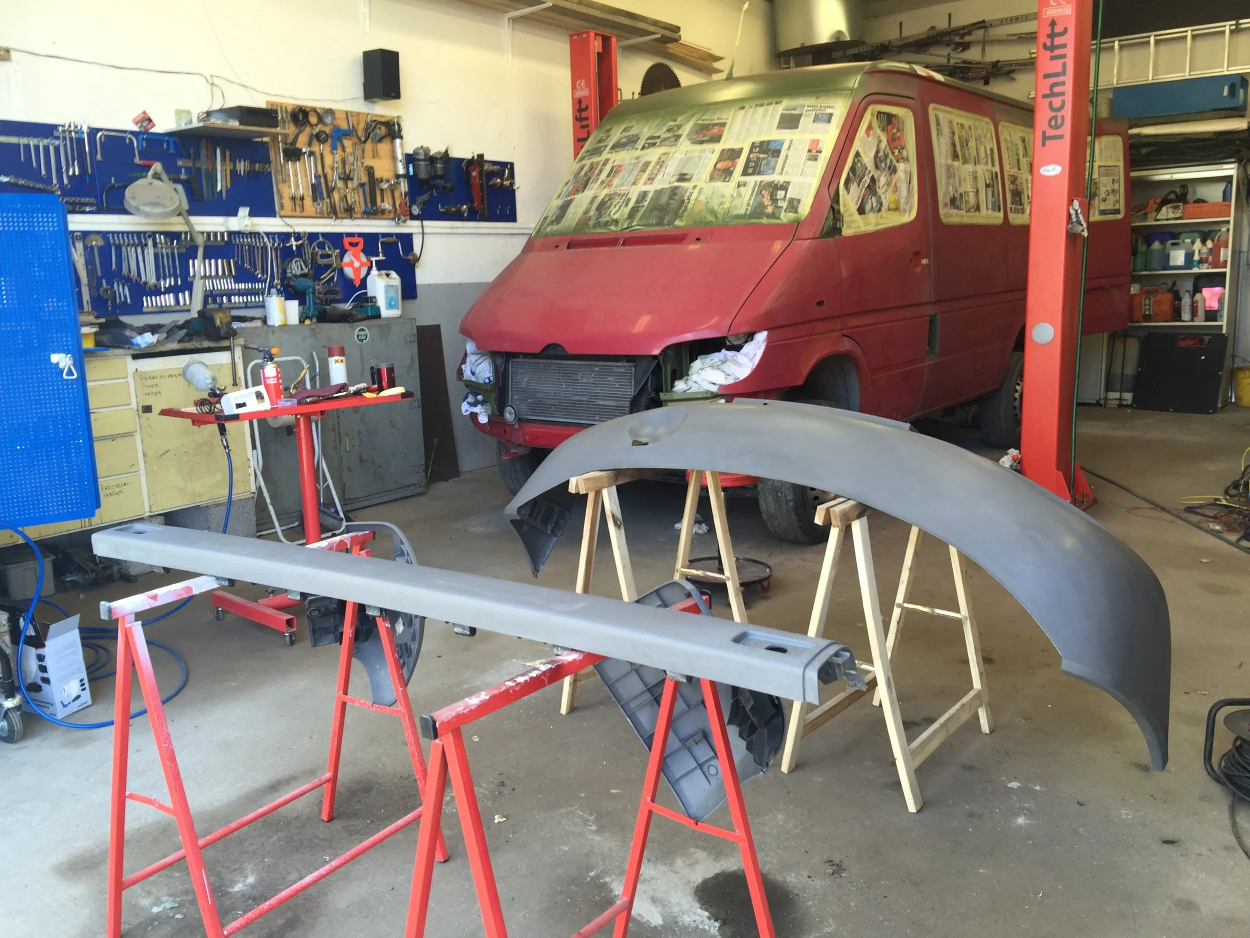 Auto body workshop with the PhotoVan being customized. The van's windshield is covered with newspaper, and a gray bumper is placed on wooden stands.