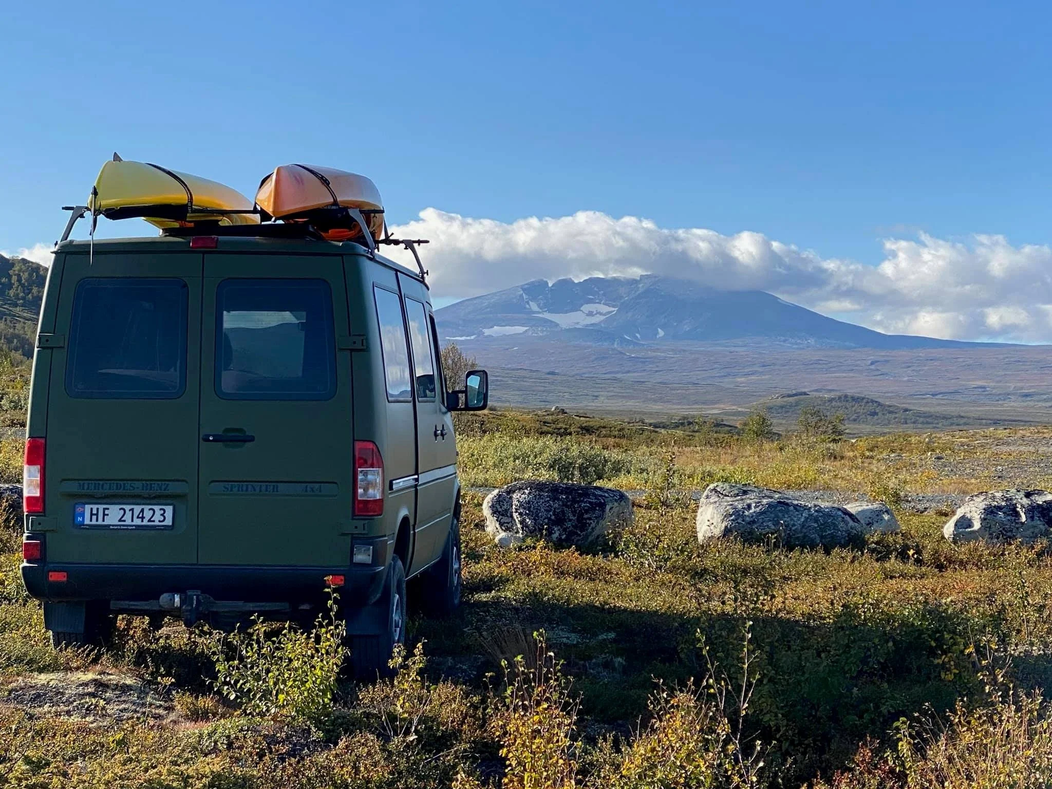 The Photovan with kayaks on the roof parked in a field with mountains in the background.