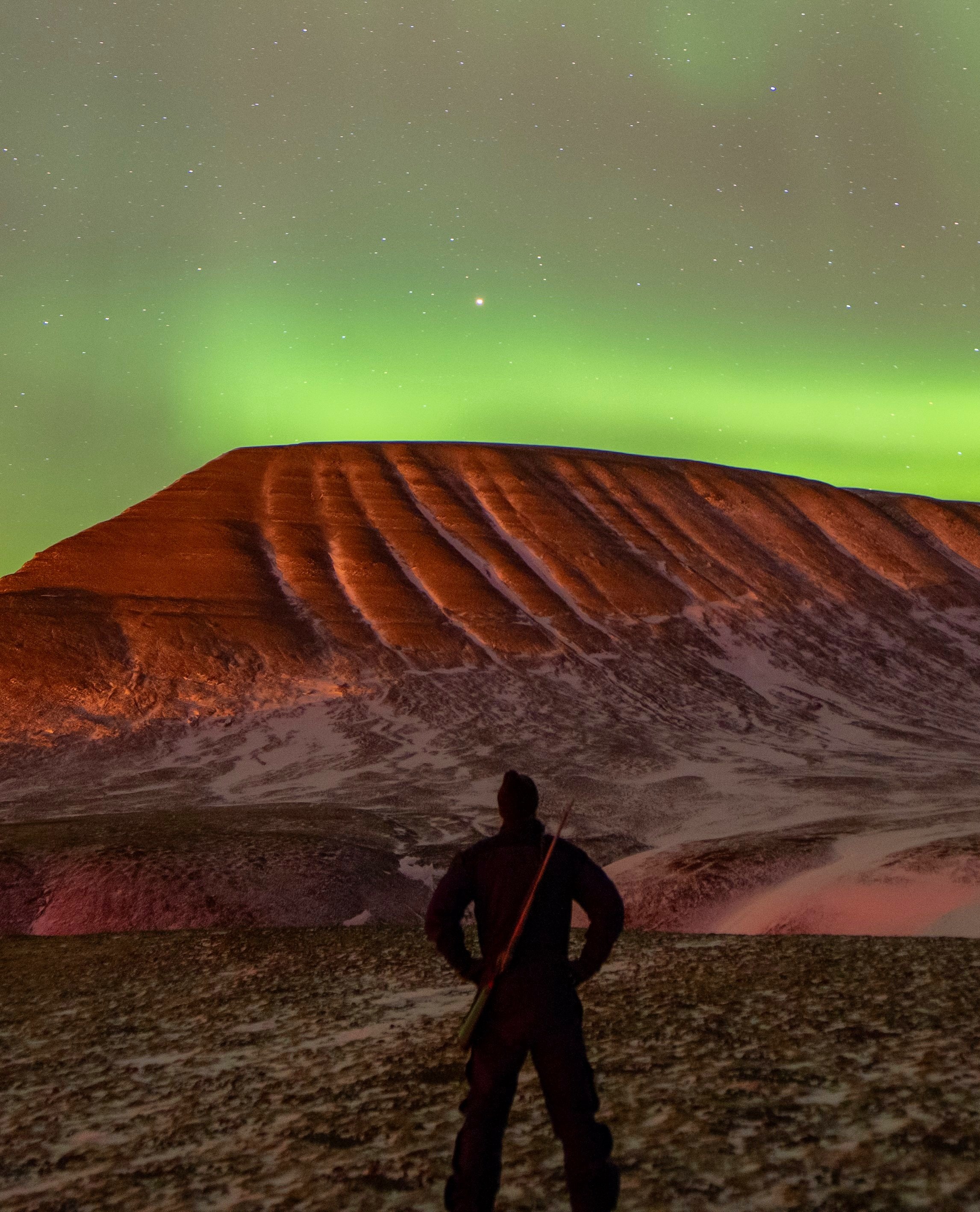 Svenn Erik Høylie standing at night, looking at aurora borealis and a snow-covered mountain in the background. He also has a rifle for polarbear protection.