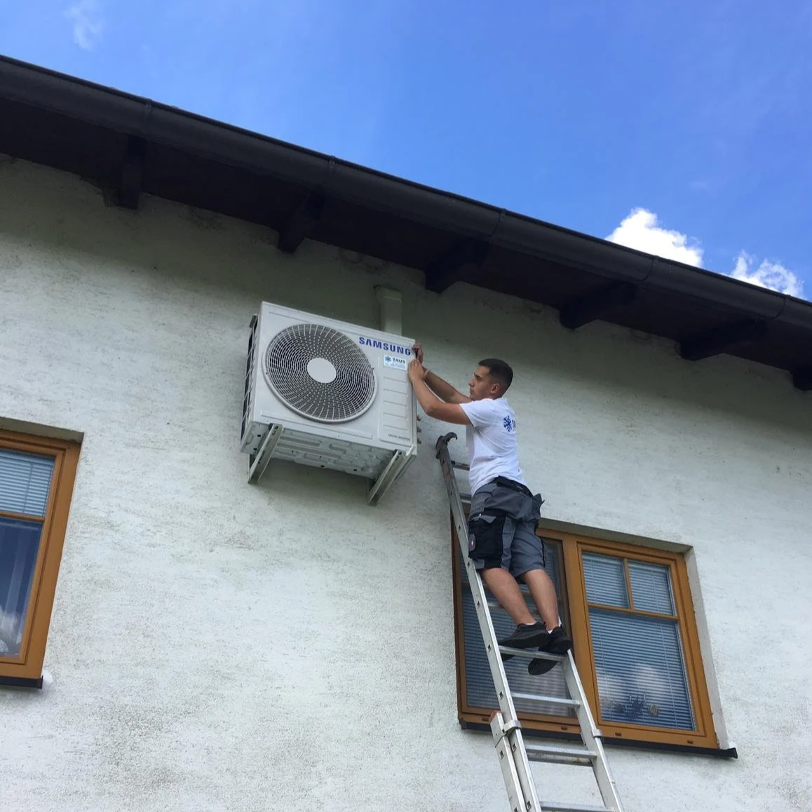 Ein Mann steht auf einer Leiter vor einem Haus und arbeitet an einer Klimaanlage, die an der Außenwand installiert ist.