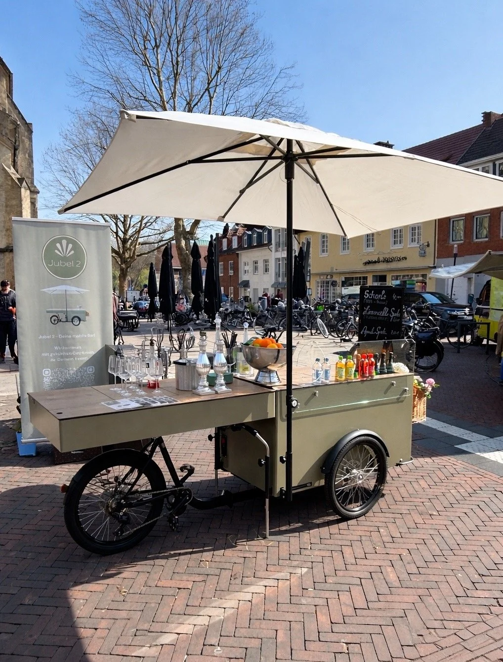 Ein mobiles Getränke- und Snack-Stand auf einem Bürgersteig mit Sonnenschirm. Im Hintergrund sind Bäume, Fahrradständer und Gebäude zu sehen, bei schönem Wetter im Freien.