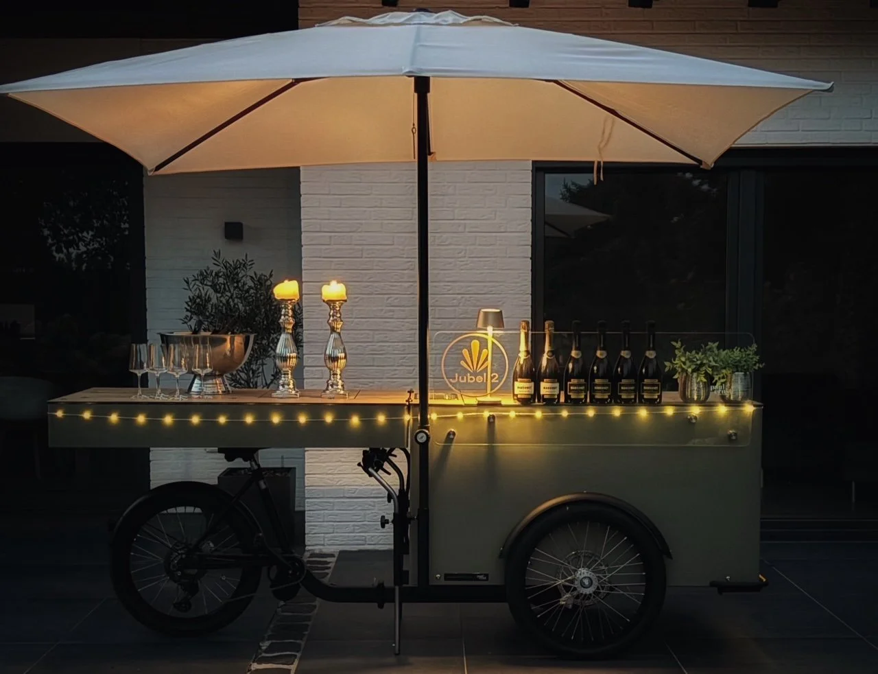 Ein Bierwagen mit dekorativen Lichtern, Flaschen und Gläsern, im Freien bei Dämmerung. Eine große weiße Sonnenschirm deckt den Wagen ab.