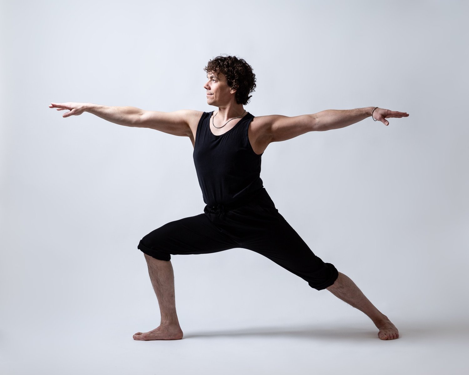 A man performing a yoga pose in a black sleeveless shirt and black pants against a plain light gray background.
