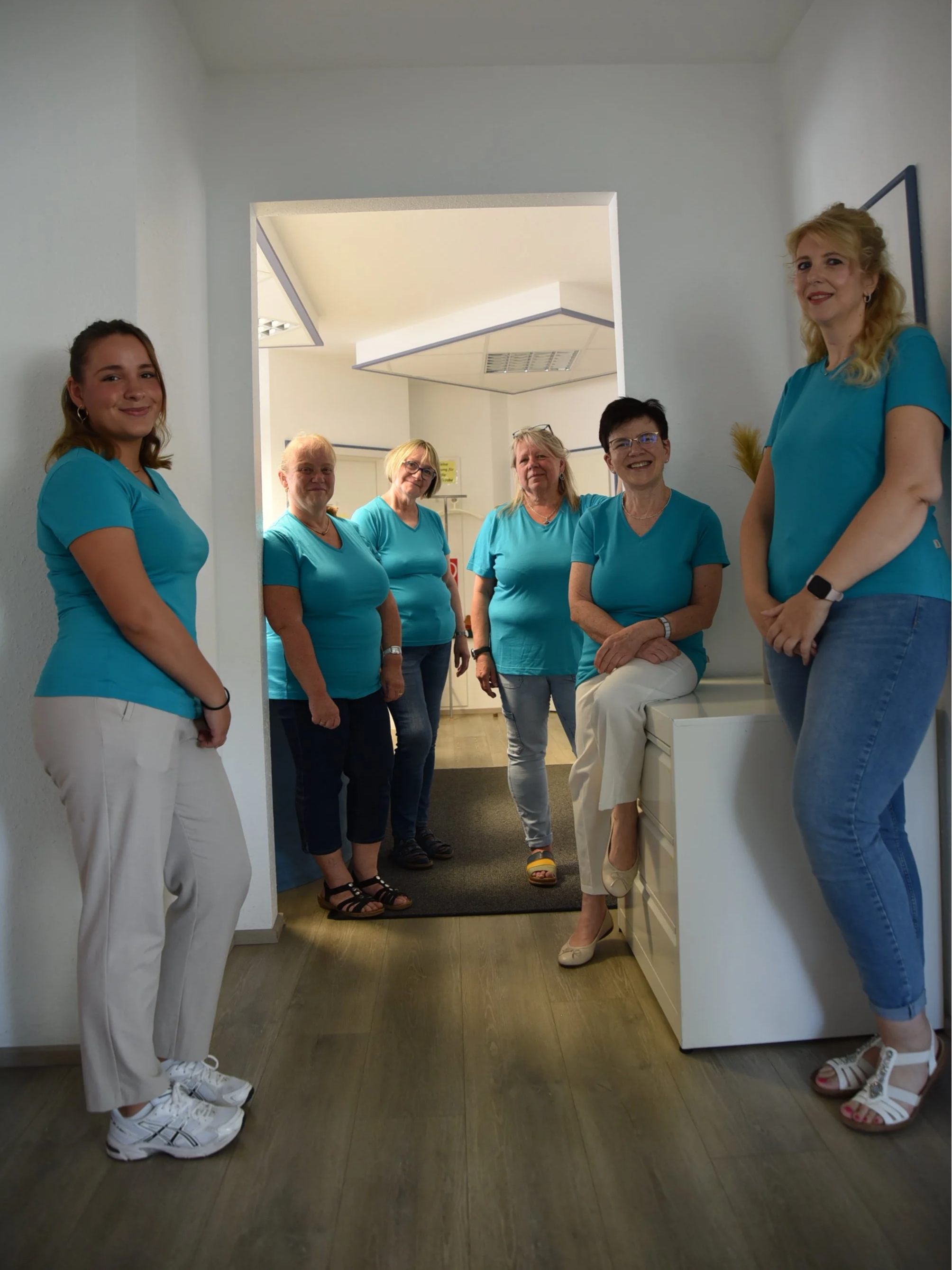 Sieben Frauen in blauen Oberteilen und weißen oder blauen Hosen stehen in einem Flur, einige lehnen an Schränken oder Wänden, alle lächeln zur Kamera.