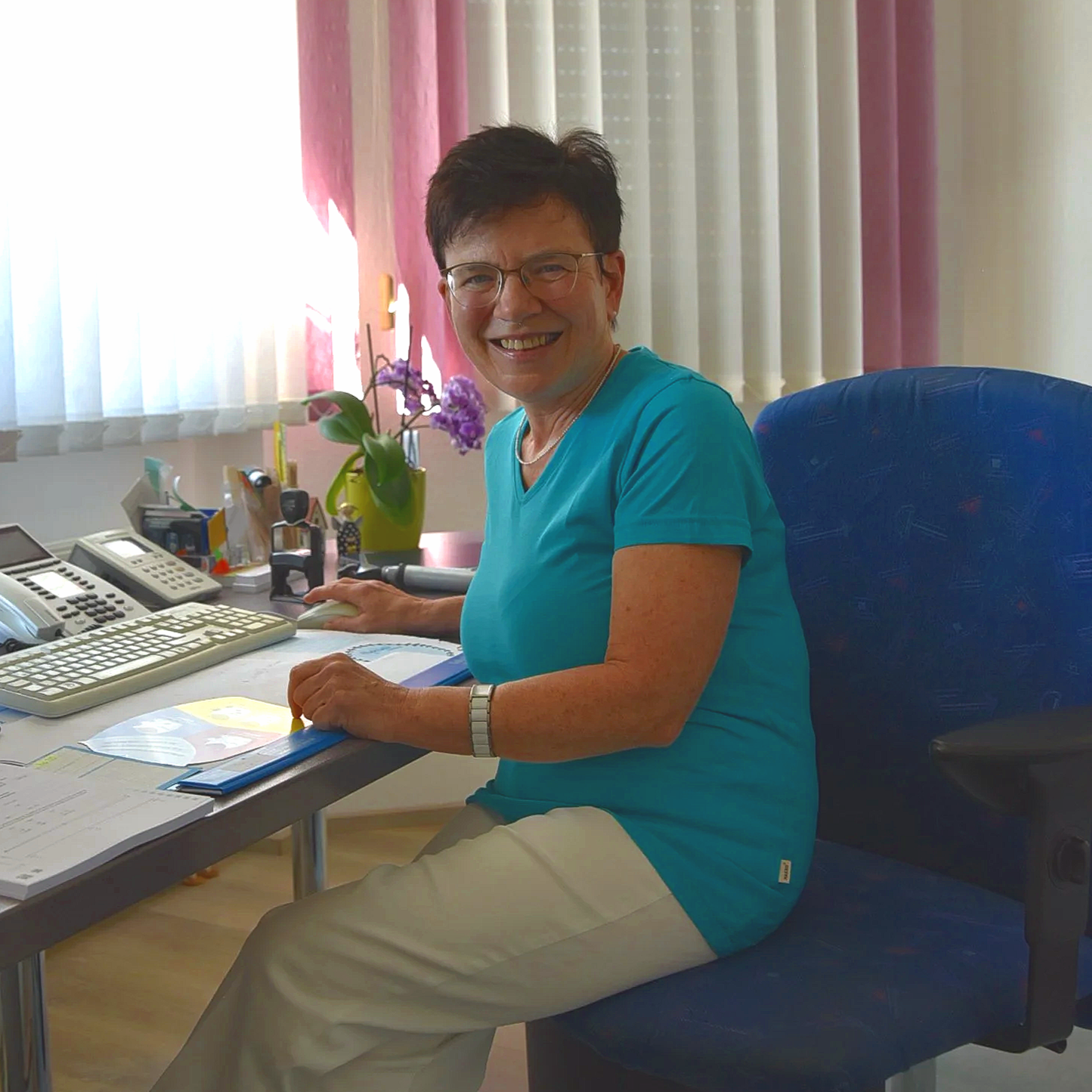 Eine lächelnde Frau sitzt an einem Schreibtisch in einem Büro, mit Papieren, einem Tastatur und Telefonen auf dem Tisch, im Hintergrund bunte Vorhänge und eine Pflanze.