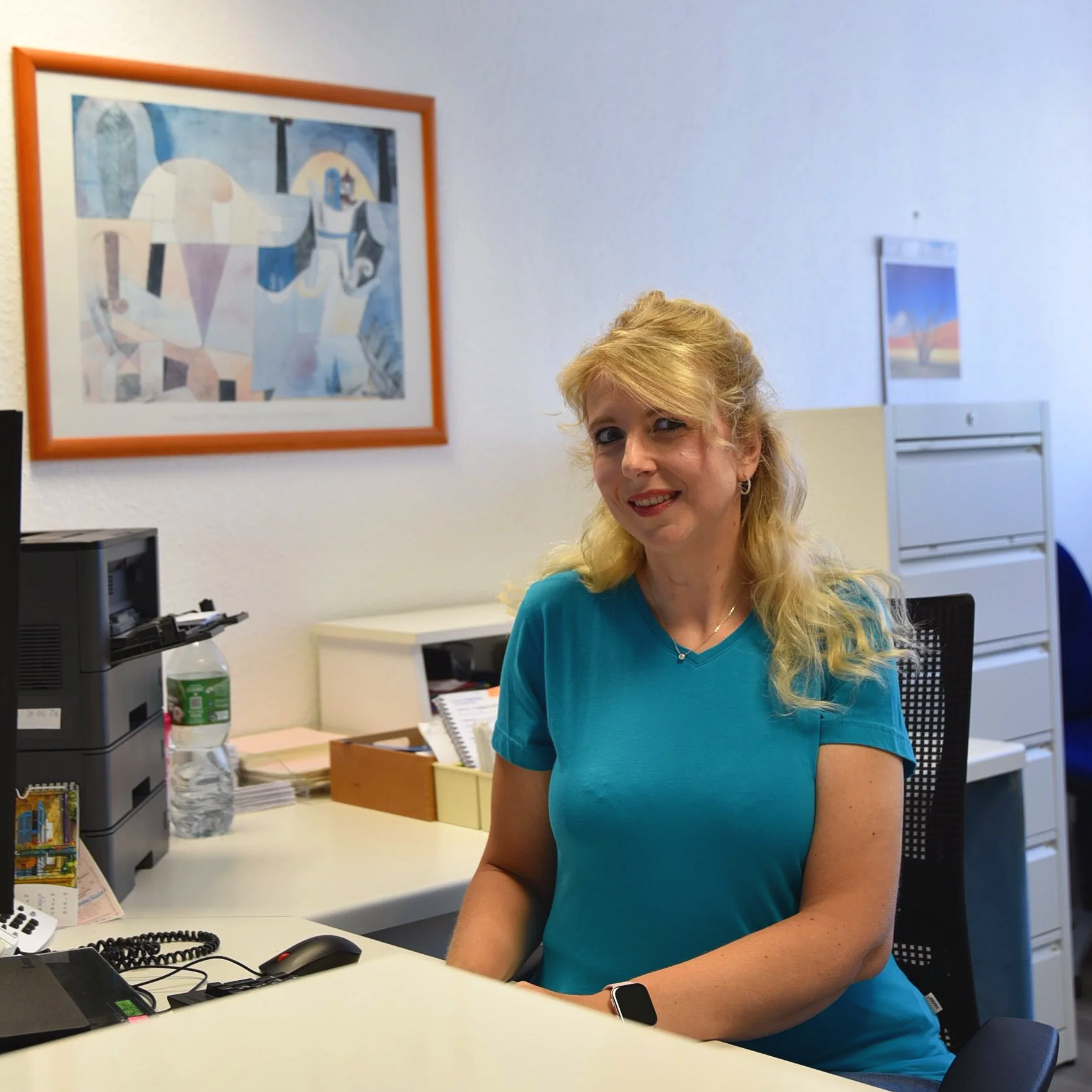 Freigestellte Frau mit blonden Haaren und blauer Bluse sitzt an einem Schreibtisch mit Computerzubehör in einem Büro, hinter ihr stehen Aktenordner und Tischkalender, Wandbild im Hintergrund.
