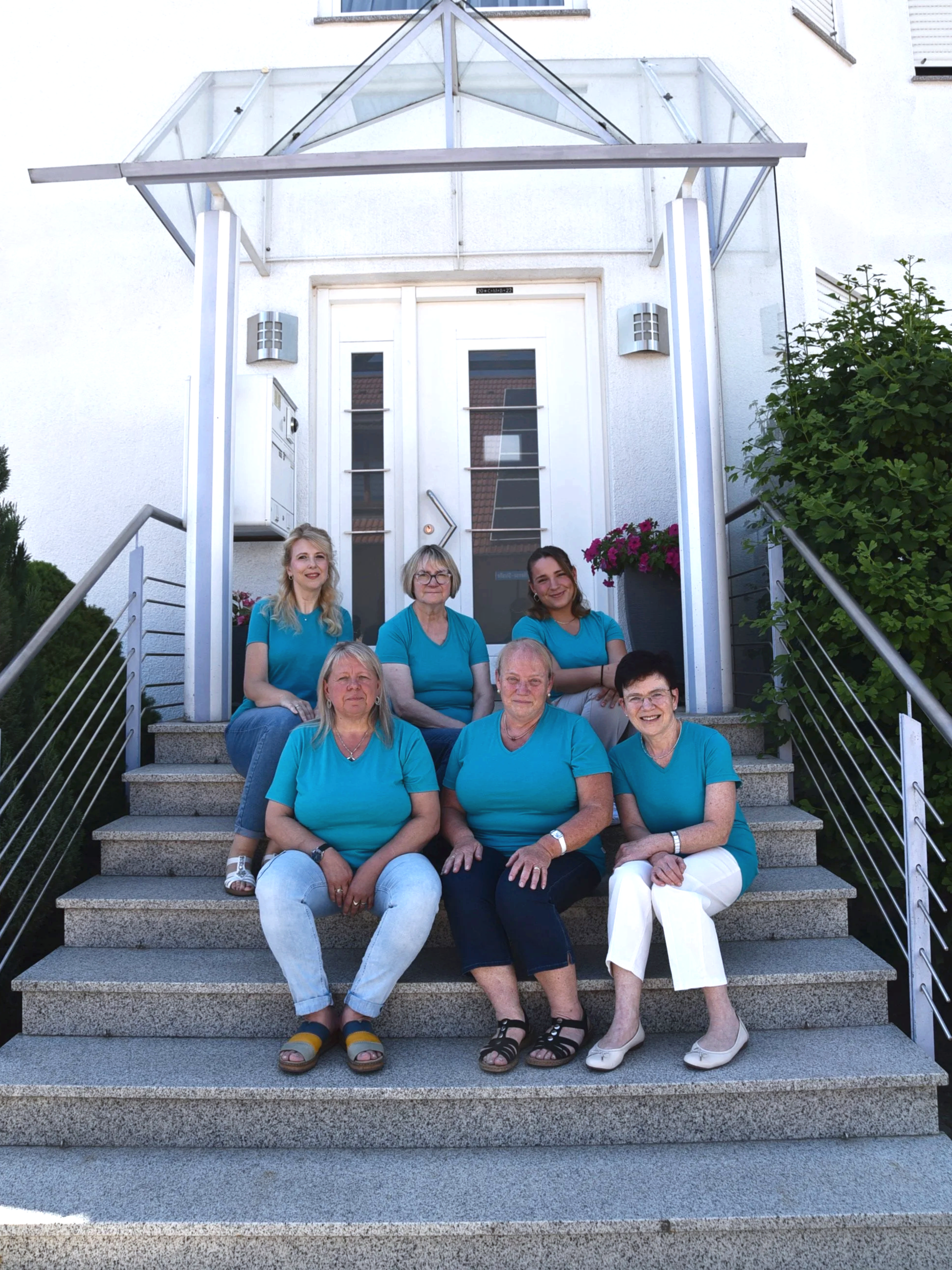 Gruppenfoto von acht Frauen sitzend und stehend auf den Steintreppen vor einem weißen Haus mit Glasüberdachung. Sie tragen gleichfarbige blaue T-Shirts, einige tragen Sandalen oder Schuhe, alle lächeln.