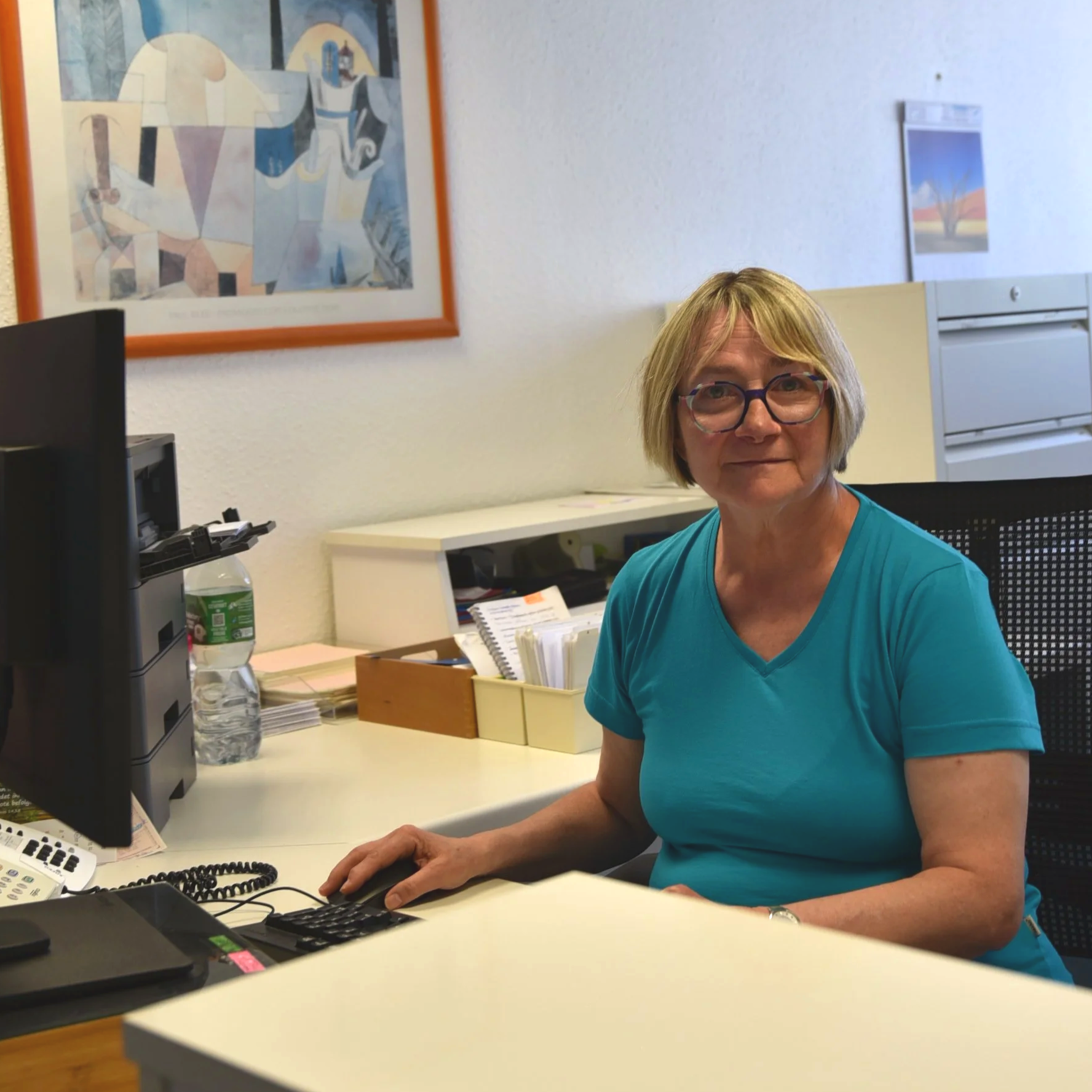 Eine ältere Frau mit kurzen grauen Haaren und Brille sitzt an einem Schreibtisch und arbeitet am Computer in einem Büro.