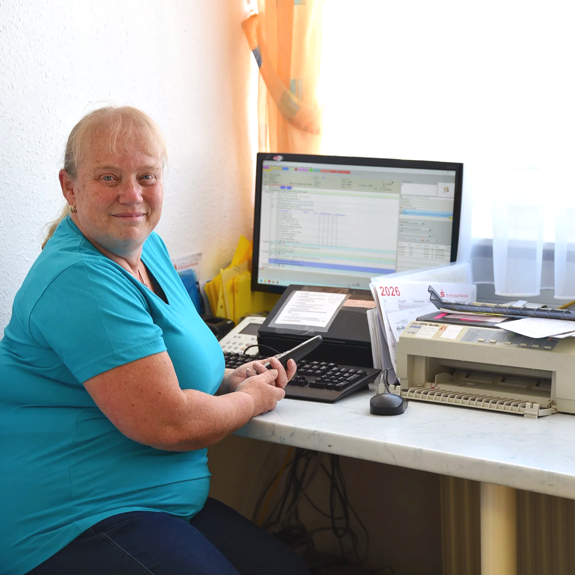 Eine ältere Frau mit blonden Haaren in blauer Kleidung sitzt an einem Schreibtisch vor einem Computerbildschirm und hält eine Fernbedienung in der Hand. Im Hintergrund befinden sich digitale Dokumente und Akten. Die Frau lächelt in die Kamera.