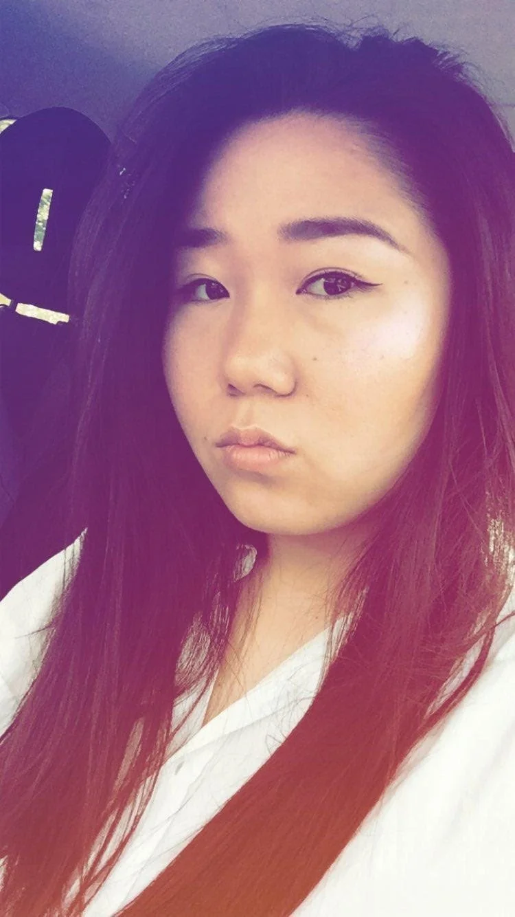 A young woman with long dark hair, in a white top, sitting in a vehicle, looking at the camera.