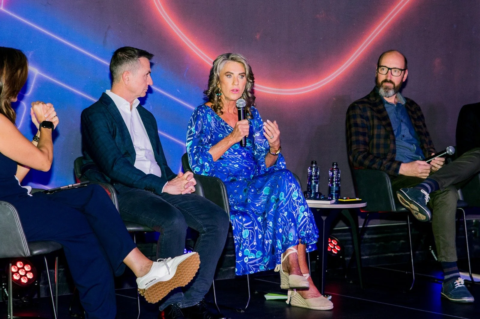 Panel of five people seated with Alison Weihe speaking into a microphone. The background features a dark space-themed design with red and blue neon lines.