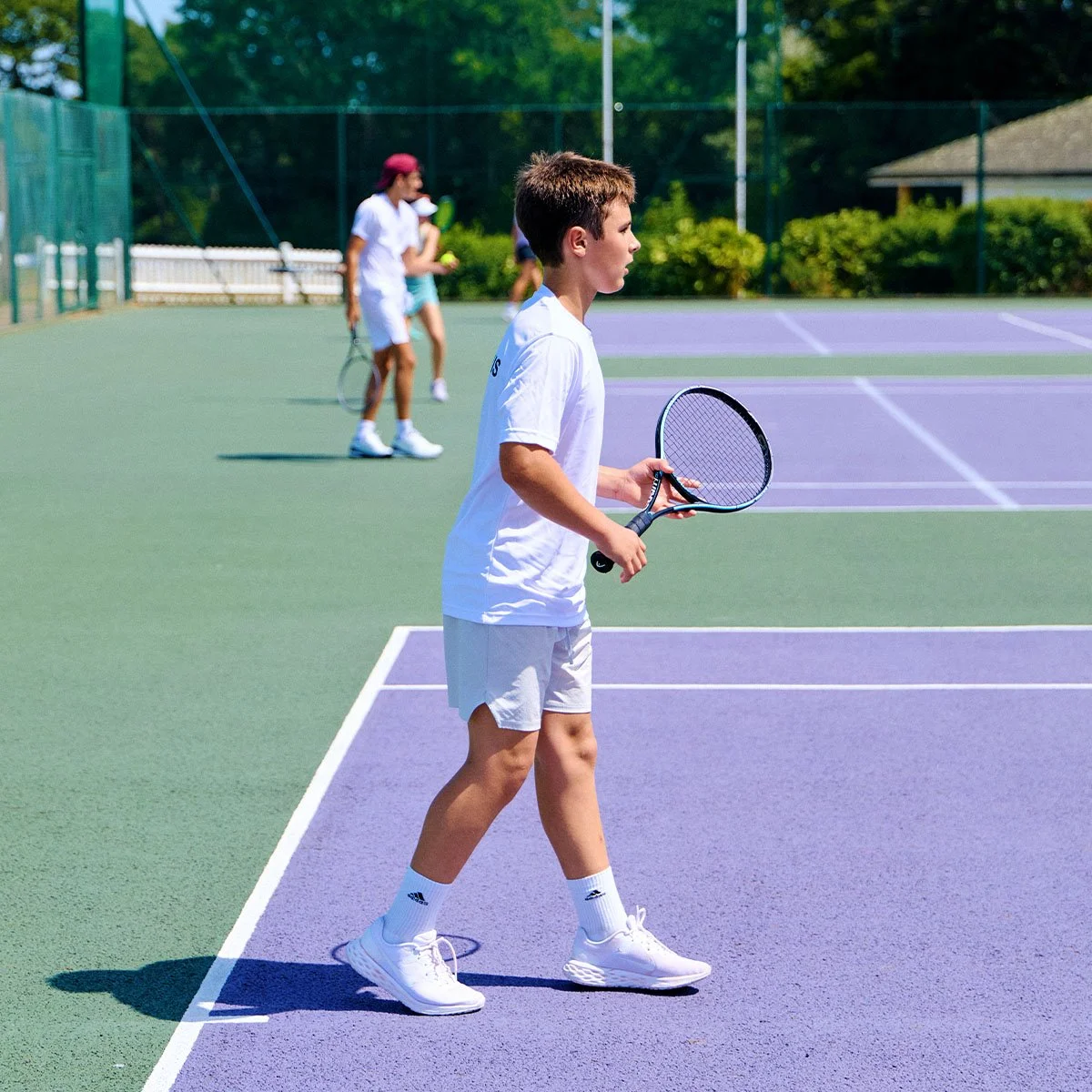 Joven tenista vestido de blanco y sujetando una raqueta en una pista de tenis exterior, con otros jugadores al fondo.