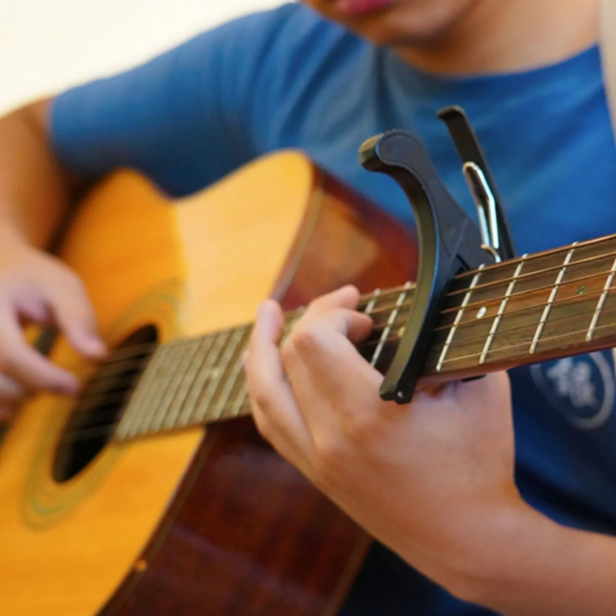 Primer plano de las manos de un joven tocando una guitarra acústica, con una cejilla colocada en el mástil.