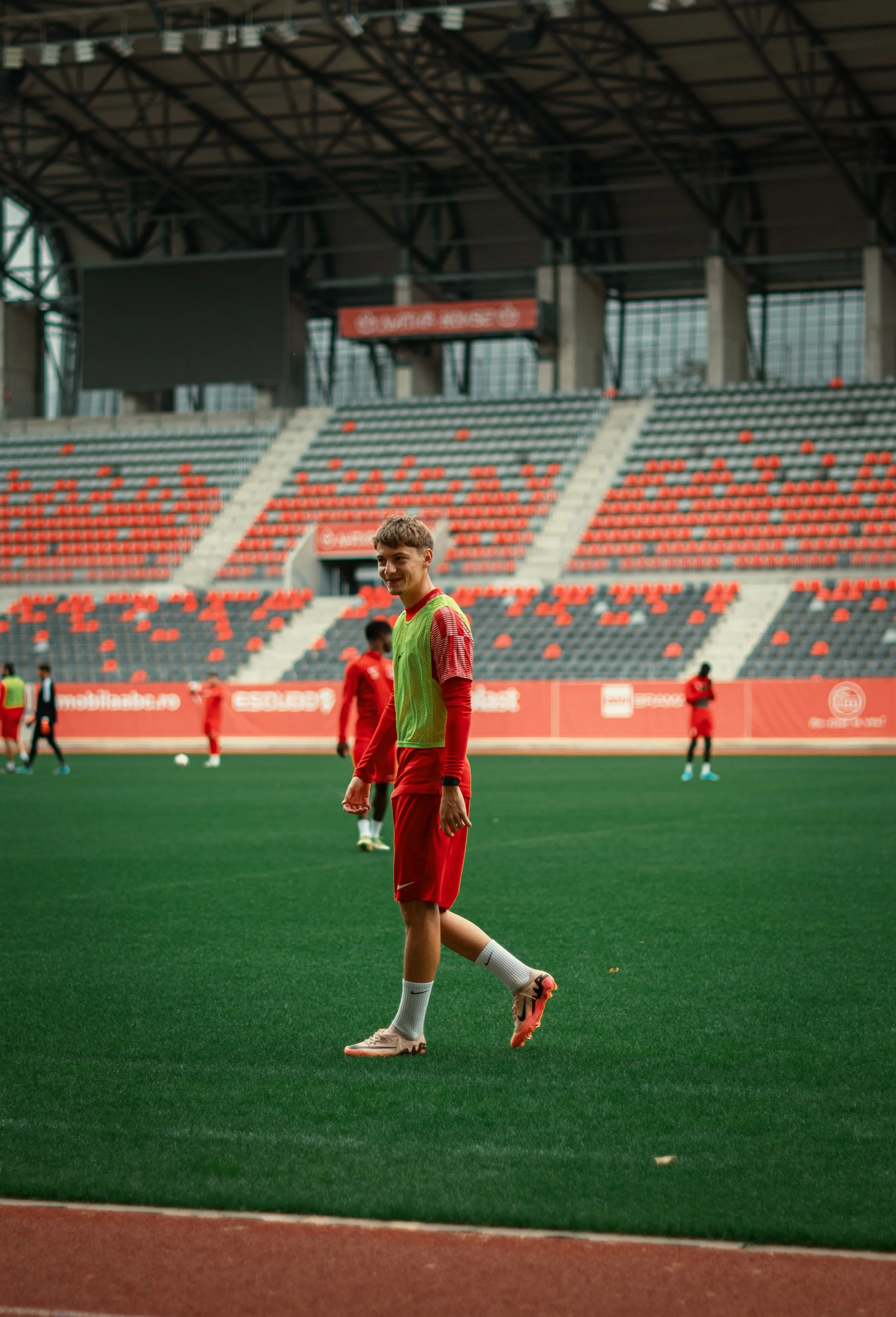 Young soccer player in red uniform and green bib on a field, with a stadium in the background.